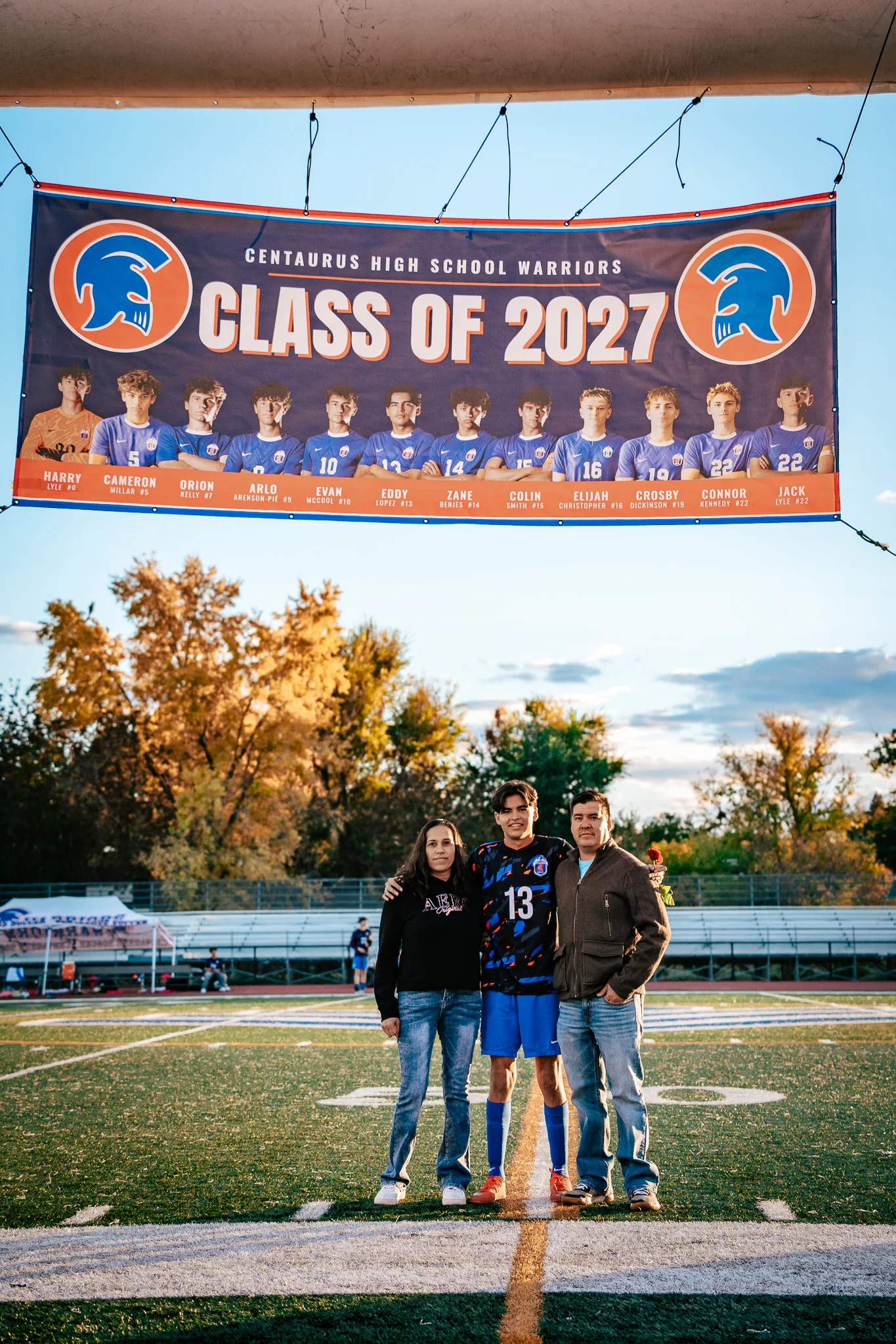 Beneath a Class of 2027 banner, Centaurus number 13 stands between a woman in a black sweatshirt and a man in a brown jacket on the field, autumn trees behind