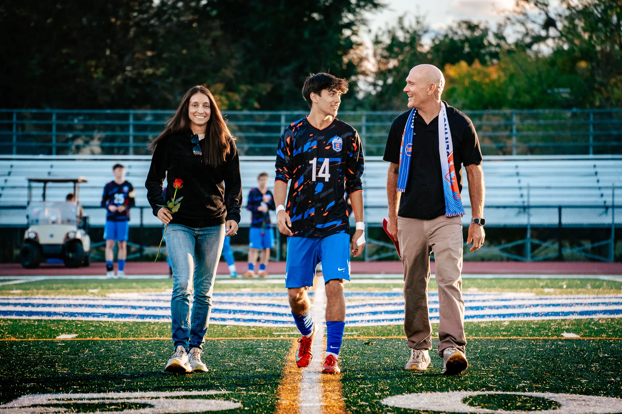 Centaurus number 14, his wrists taped, walks onto the field between a woman holding a red rose and a bald man in a blue and orange scarf who laughs as he looks at him, a golf cart on the sideline behind