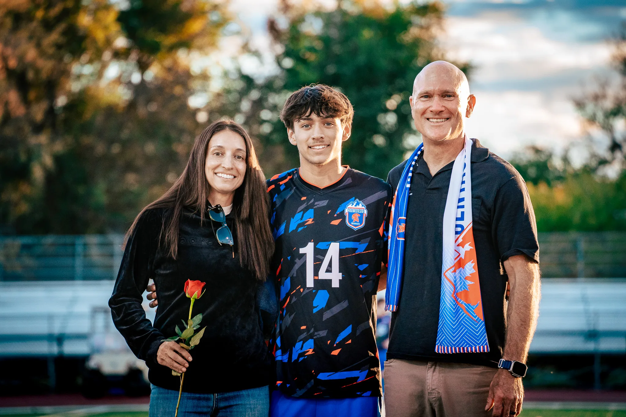 Close up, Centaurus number 14 smiles beside a woman in a black fleece holding a red rose and a bald man in a black polo and blue and orange scarf, trees blurred behind