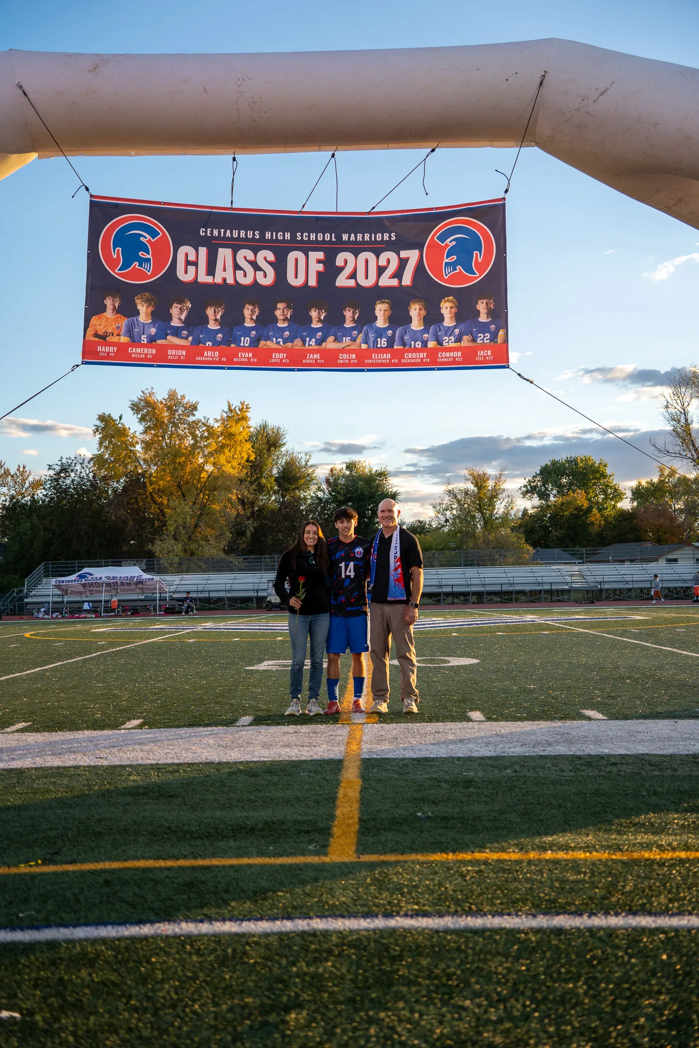 Under a Class of 2027 banner hung from an inflatable arch, Centaurus number 14 stands on the field with a woman holding a red rose and a bald man in a blue and orange scarf, autumn trees and bleachers behind