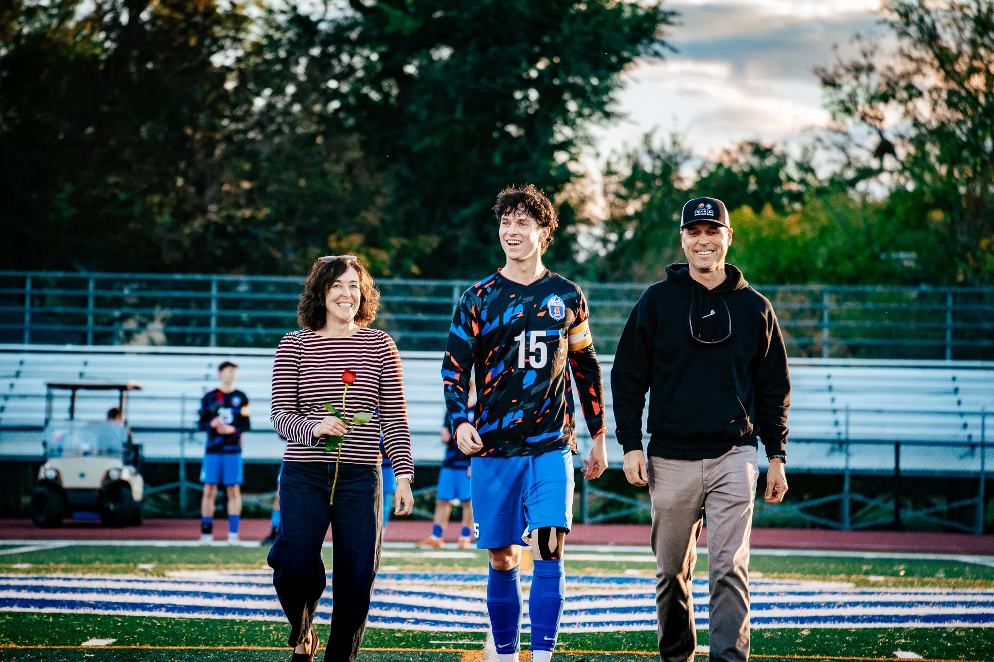 Centaurus number 15, wearing a captain's armband, laughs as he walks onto the field between a woman in a striped top holding a red rose and a man in a black cap and hoodie, a golf cart behind