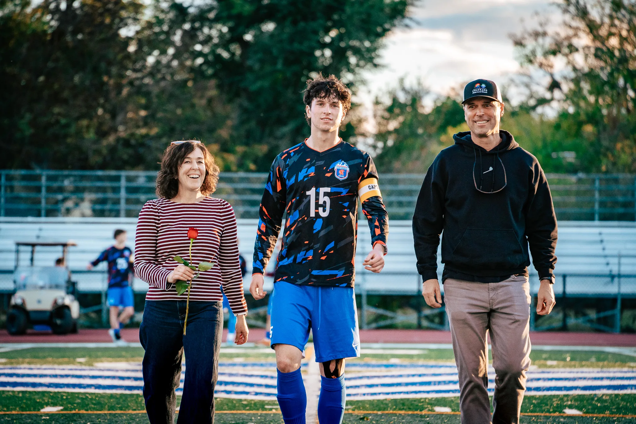 Centaurus number 15 in a captain's armband walks toward the camera between a smiling woman in a striped top holding a red rose and a man in a black cap and hoodie
