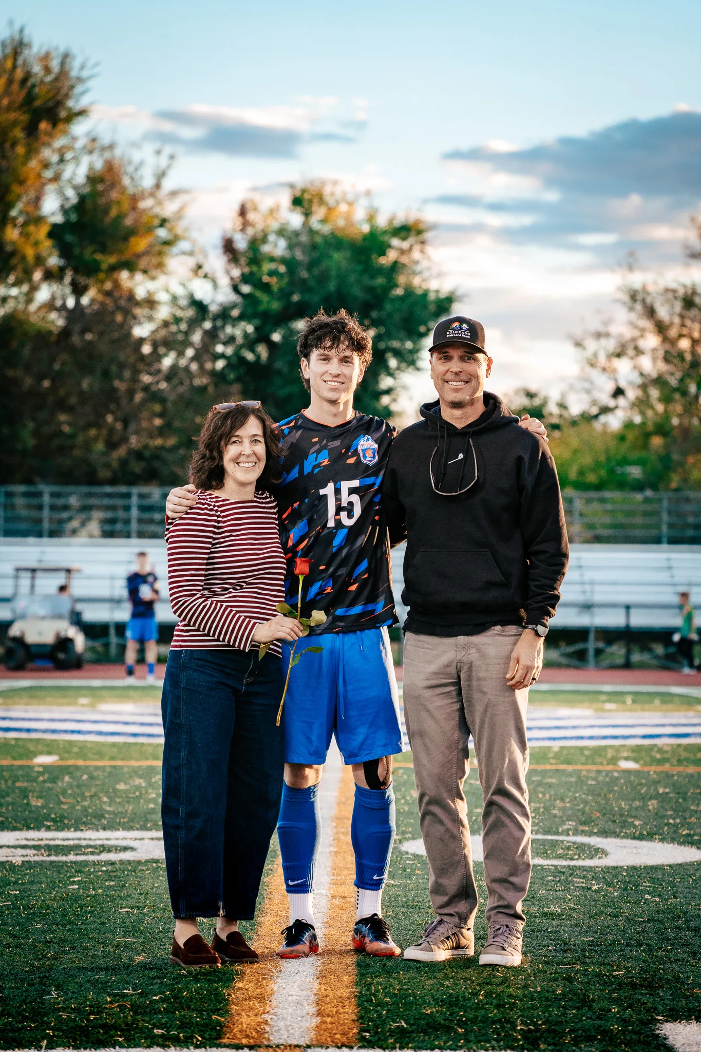 Centaurus number 15 poses at midfield between a woman in a striped top holding a red rose and a man in a black cap and hoodie, a golf cart and teammates along the sideline behind