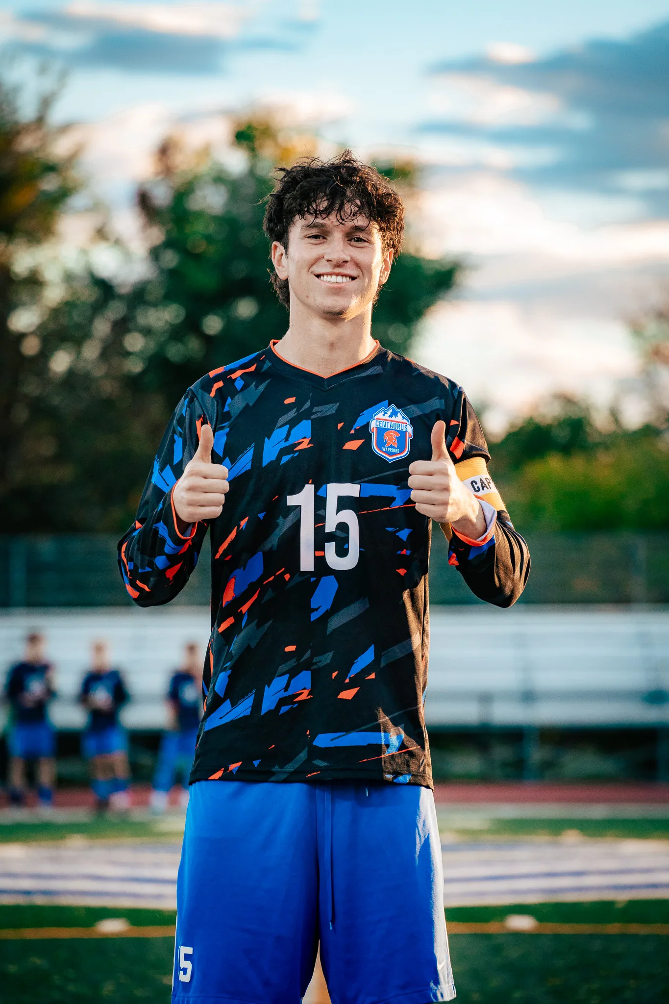 Centaurus number 15 smiles at the camera with both thumbs up, a captain's armband on his sleeve, the bleachers and trees blurred behind