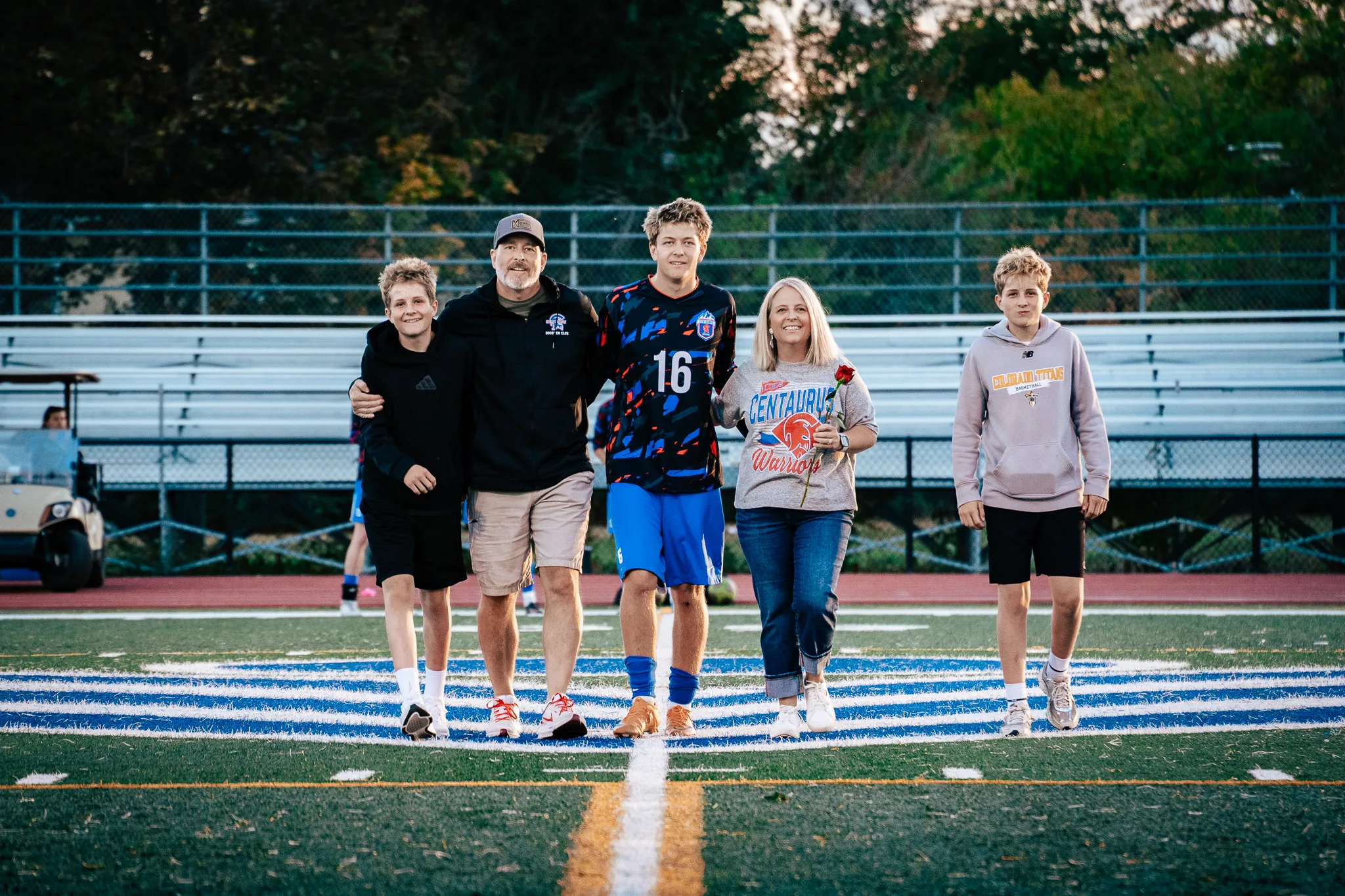 Centaurus number 16 walks onto the field arm in arm with a bearded man in a cap and a woman in a Centaurus Warriors shirt holding a red rose, a younger boy in a hoodie at each end of the line