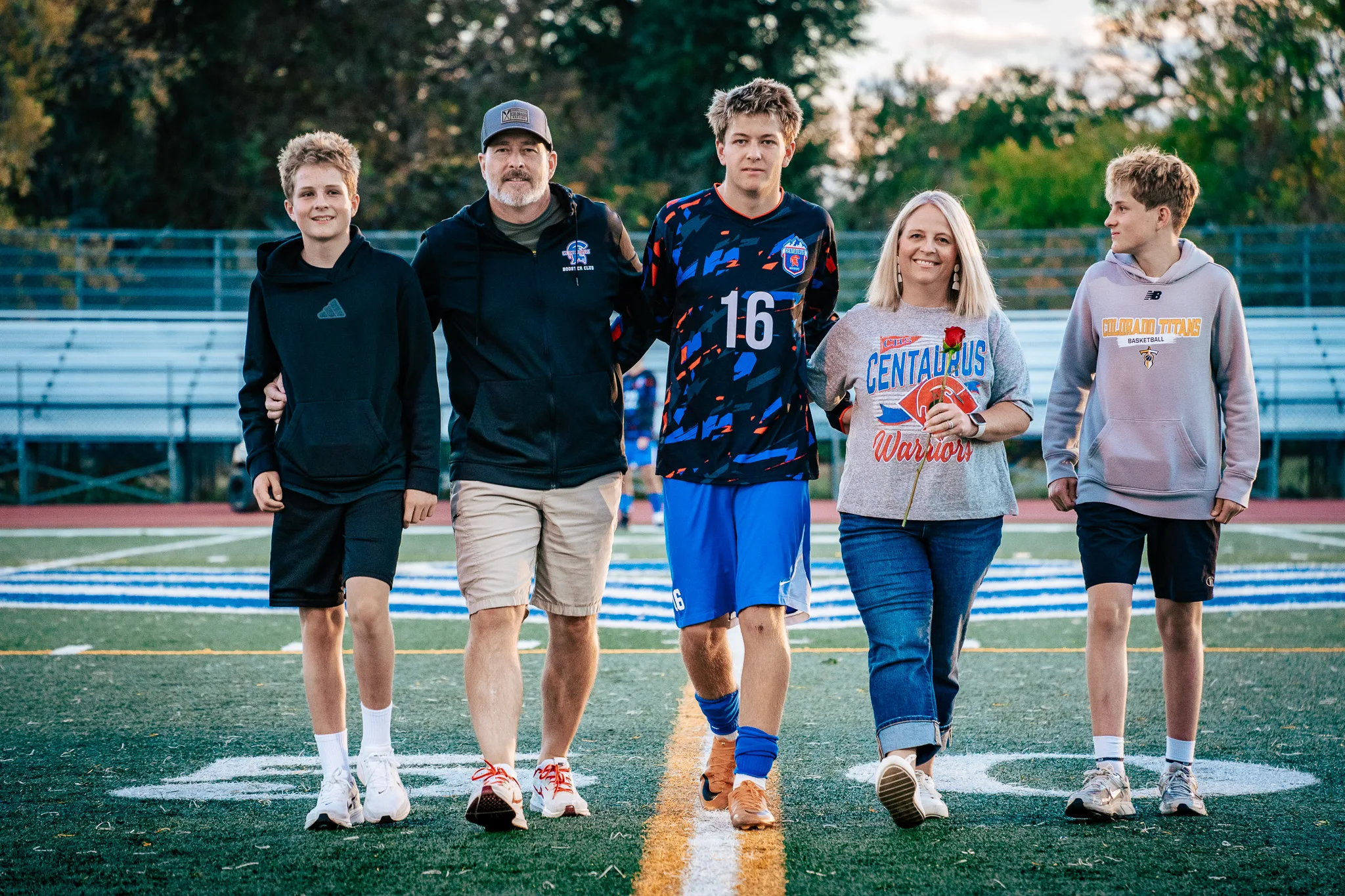Centaurus number 16 walks across the midfield logo with a bearded man in a cap, a woman in a Centaurus Warriors shirt holding a red rose and two younger boys in hoodies, all five in a line