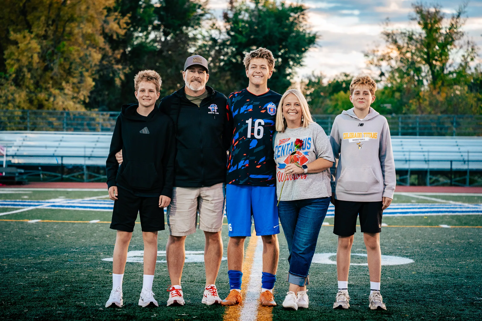 Centaurus number 16 poses at midfield in a line with a bearded man in a cap, a woman in a Centaurus Warriors shirt holding a red rose and two younger boys in hoodies, autumn trees behind