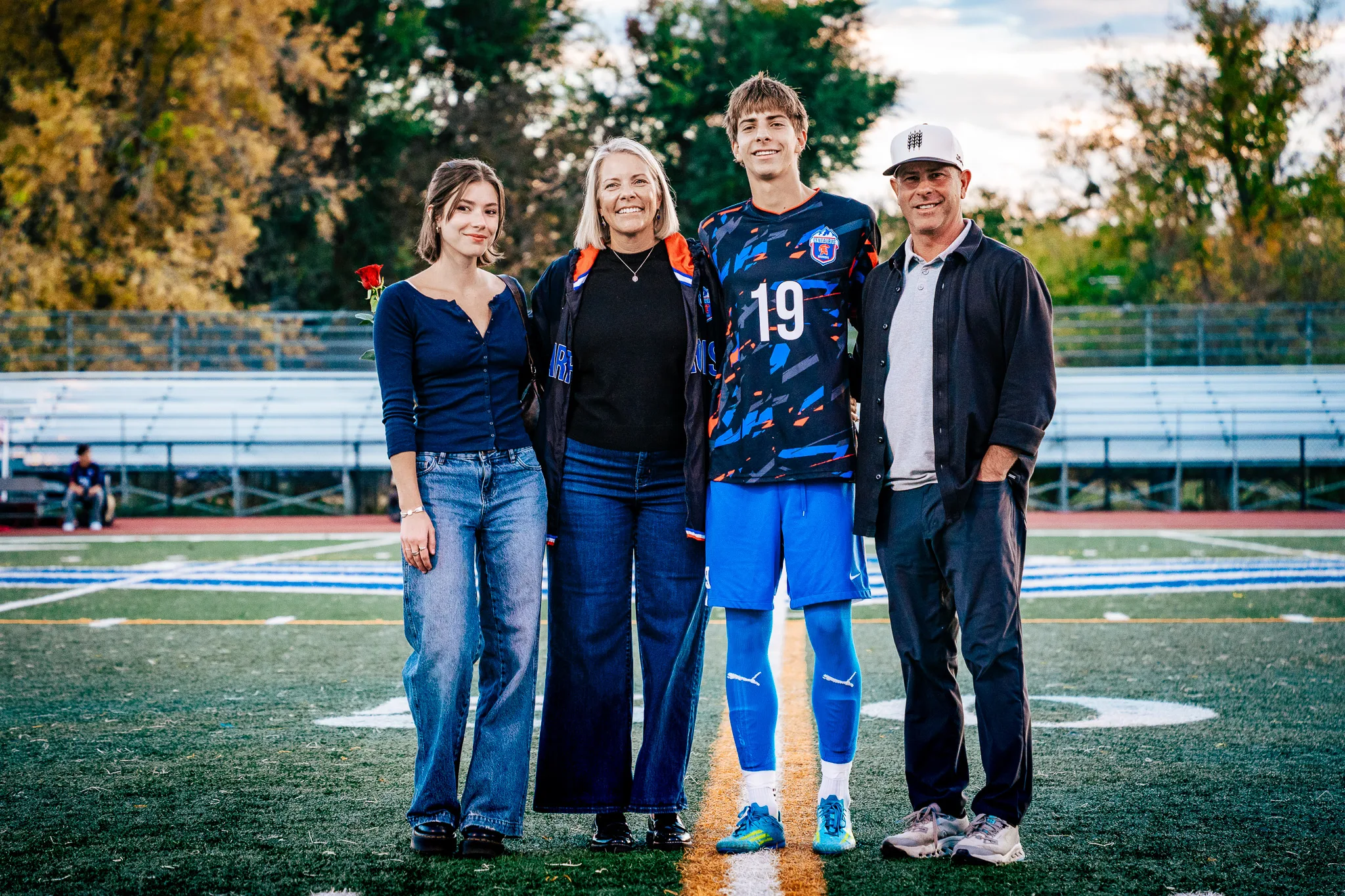 Centaurus number 19 poses at midfield with a young woman in a navy top holding a red rose, a woman in a black top and a blue and orange jacket and a man in a white cap and dark shirt, autumn trees behind