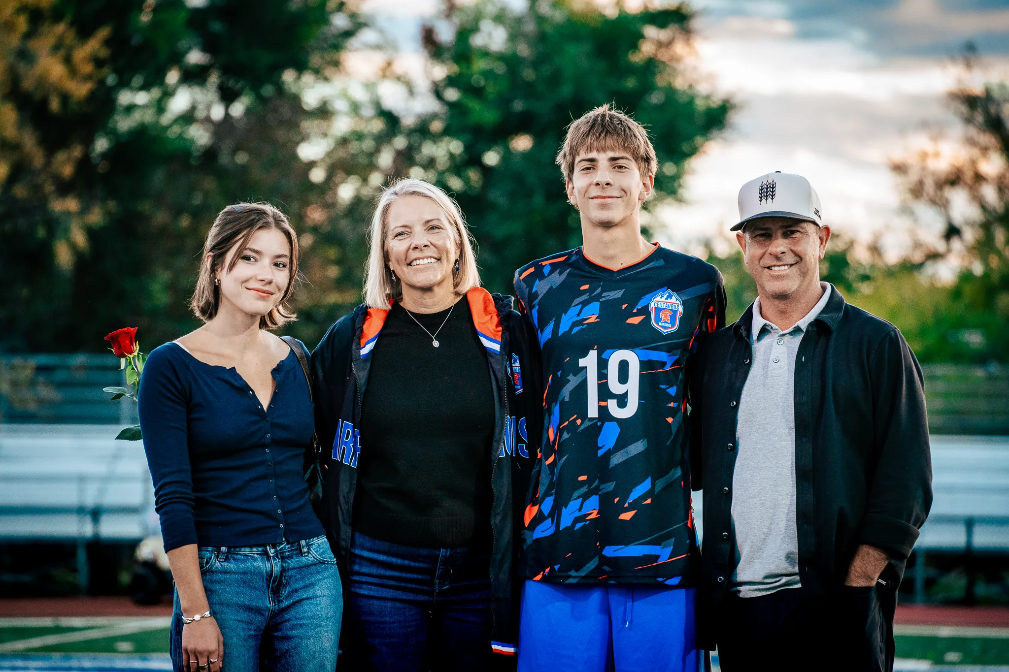 Close up, Centaurus number 19 smiles beside a young woman in a navy top with a red rose, a woman in a blue and orange jacket and a man in a white cap