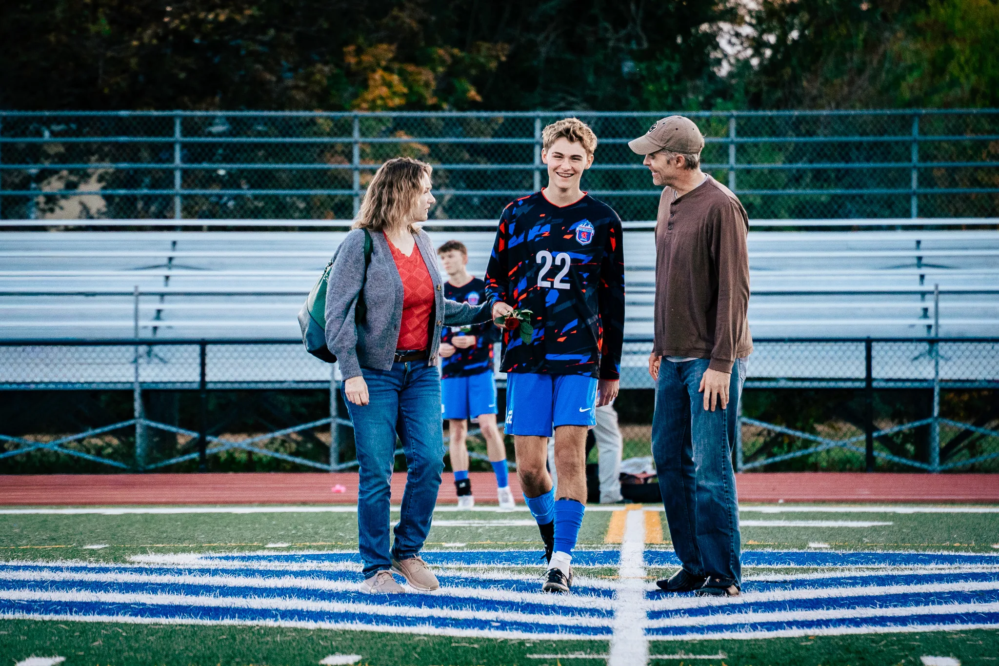 Centaurus number 22 smiles as he walks onto the field between a woman in a gray cardigan holding a red rose and a man in a brown henley and cap, a teammate waiting behind them