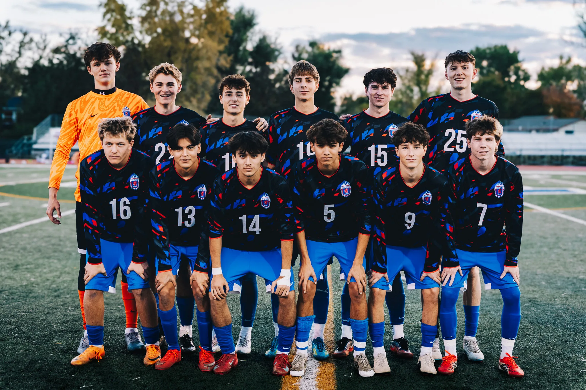 Twelve Centaurus seniors in dark patterned jerseys pose in two rows, the goalkeeper in orange at the back left and numbers 16, 13, 14, 5, 9 and 7 leaning in at the front, trees and a pale evening sky behind
