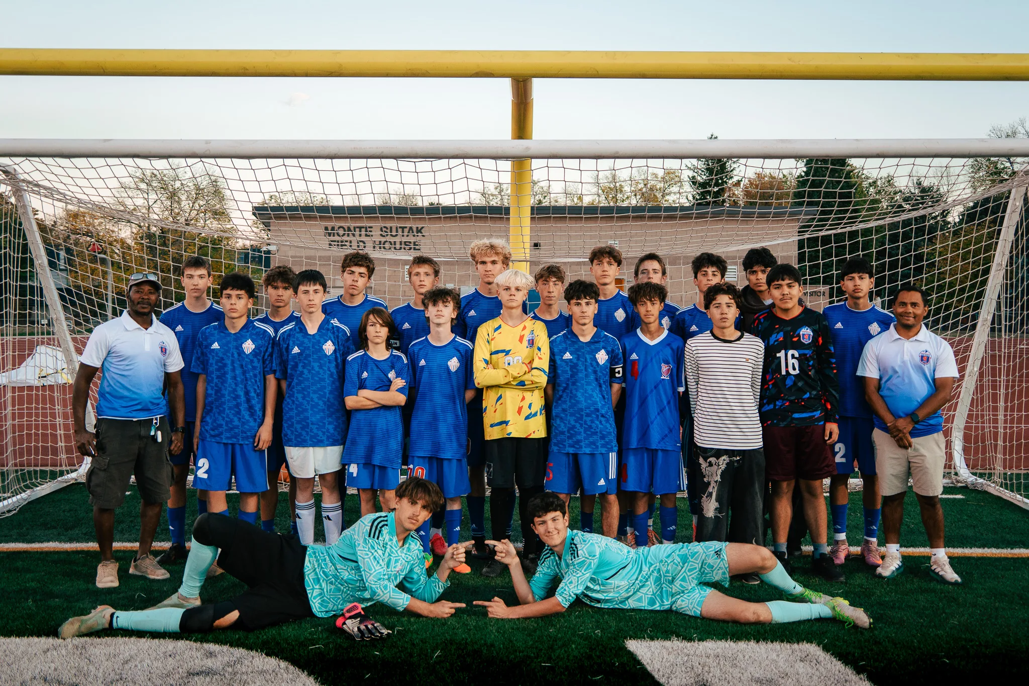 A squad in royal blue jerseys stands in rows inside a goal with two coaches in white and blue polos, a goalkeeper in yellow in the middle and two goalkeepers in teal lying on the turf in front, pointing at each other, a field house behind the net