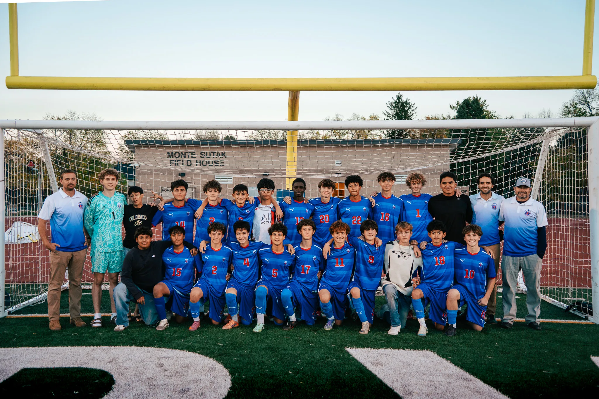 Players in royal blue jerseys with red numbers pose in front of a goal, one row kneeling and one standing, with coaches in light blue polos at either end, a goalkeeper in teal at the left, the goalposts and a clear evening sky behind