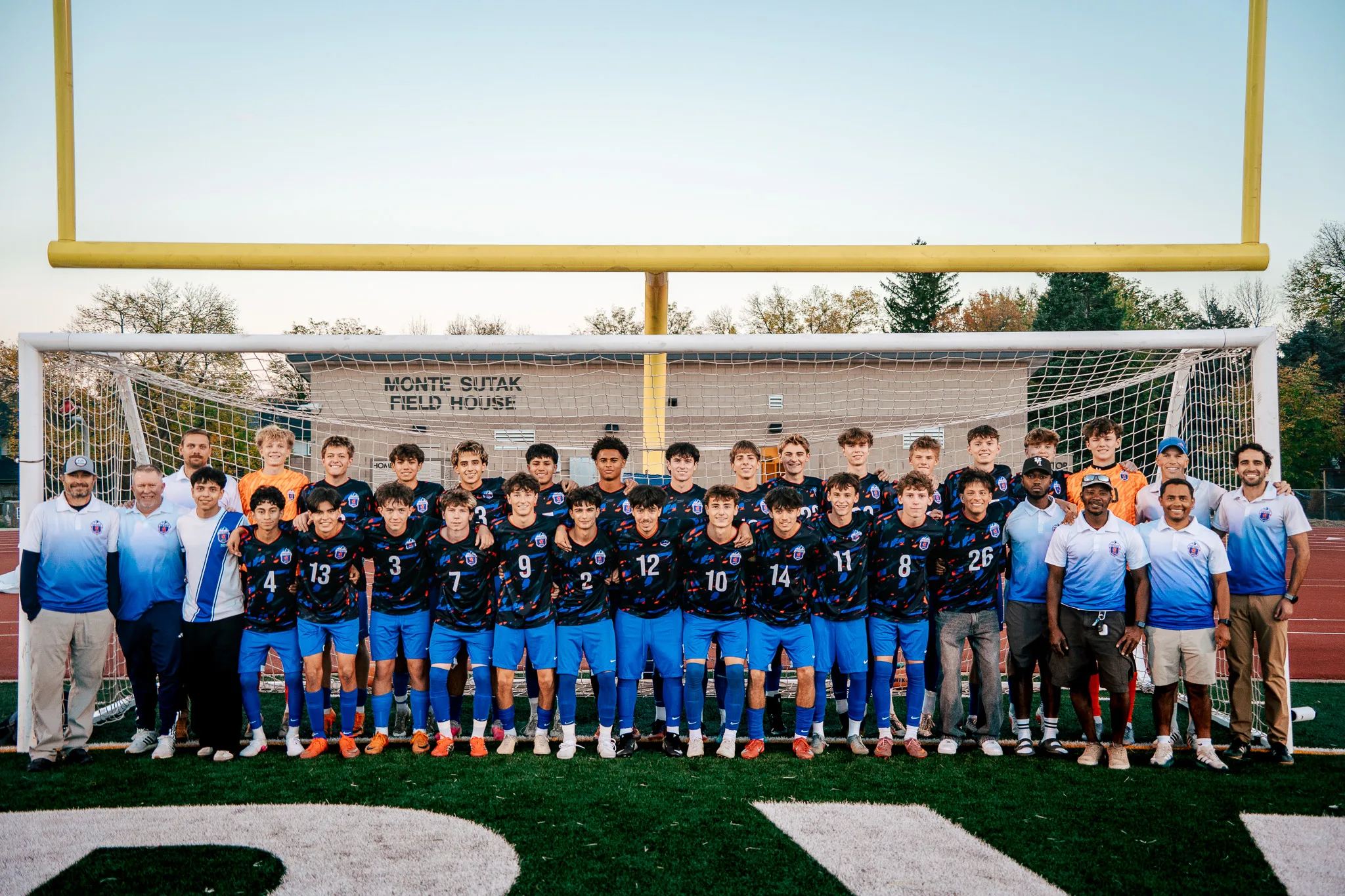The Centaurus squad in dark patterned jerseys stands in two rows in front of a goal between coaches in light blue polos, the goalkeepers in orange at the back and numbers 4, 13, 3, 7, 9, 2, 12, 10, 14, 11, 8 and 26 across the front, yellow goalposts and a pale sky behind