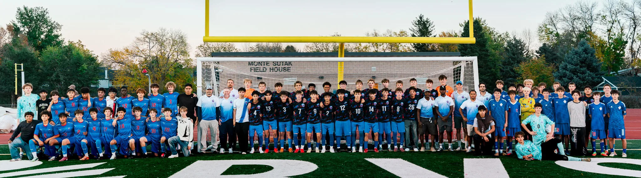 A panorama of several squads lined up in front of the goal, the Centaurus squad in dark patterned jerseys and its coaches in the middle and players in royal blue jerseys on either side, goalkeepers in teal and yellow among them, autumn trees and a field house behind