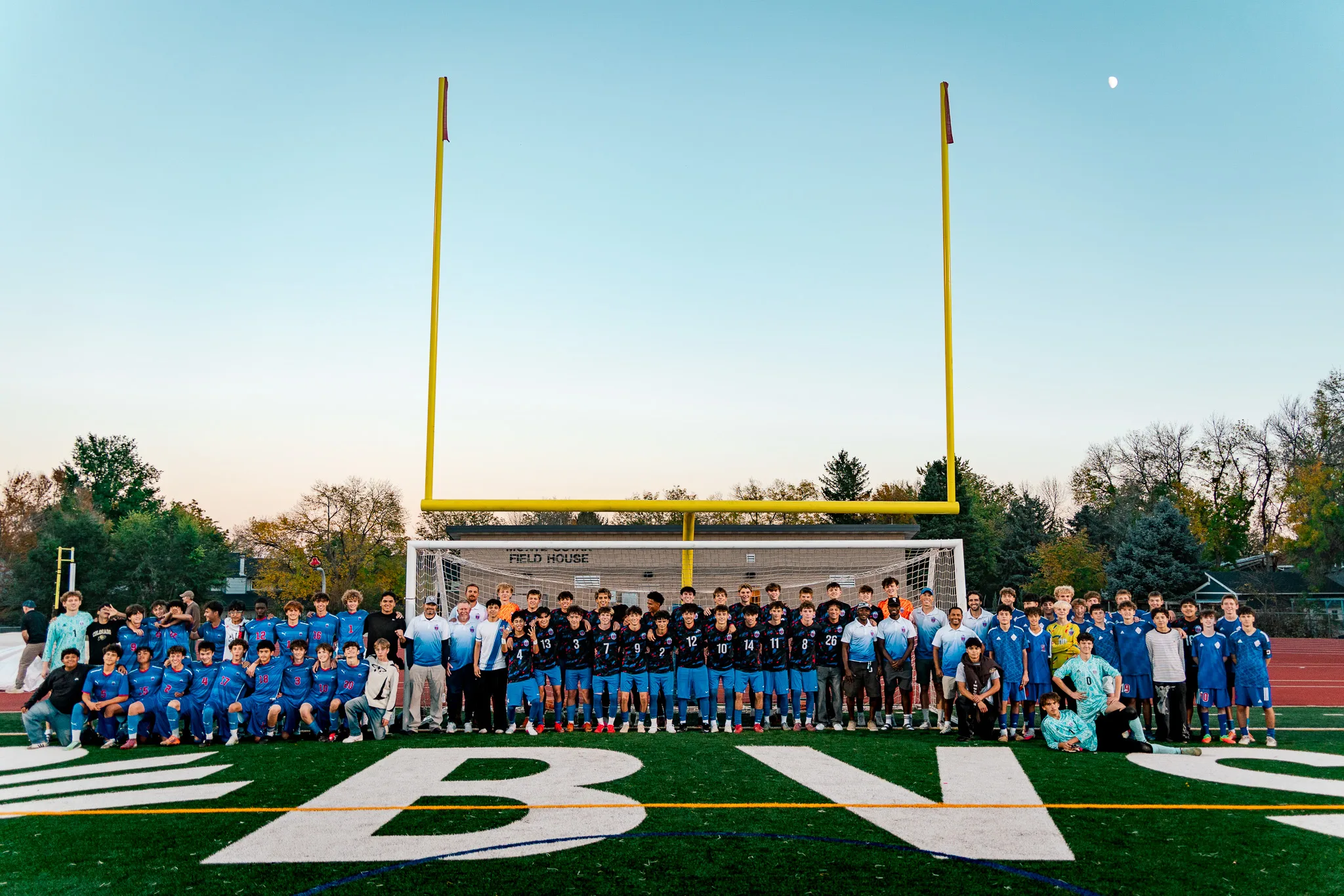 A wide view of the same lineup in front of the goal, small beneath the tall yellow goalposts, with a clear evening sky and the moon at the upper right and painted lettering on the turf in the foreground
