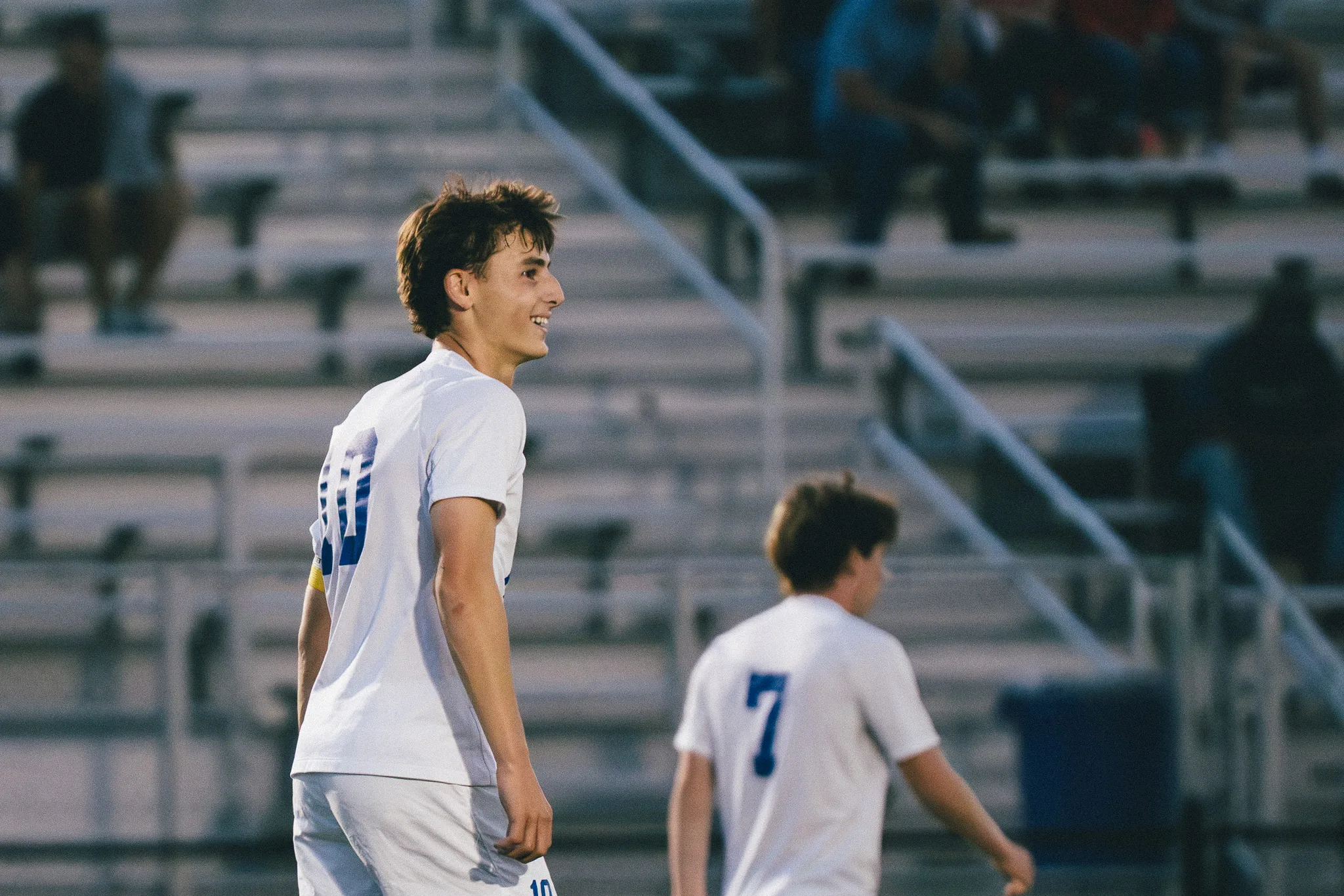 Smiling, a Centaurus player looks off to the side as teammate number 7 walks behind him, the bleachers soft in the background
