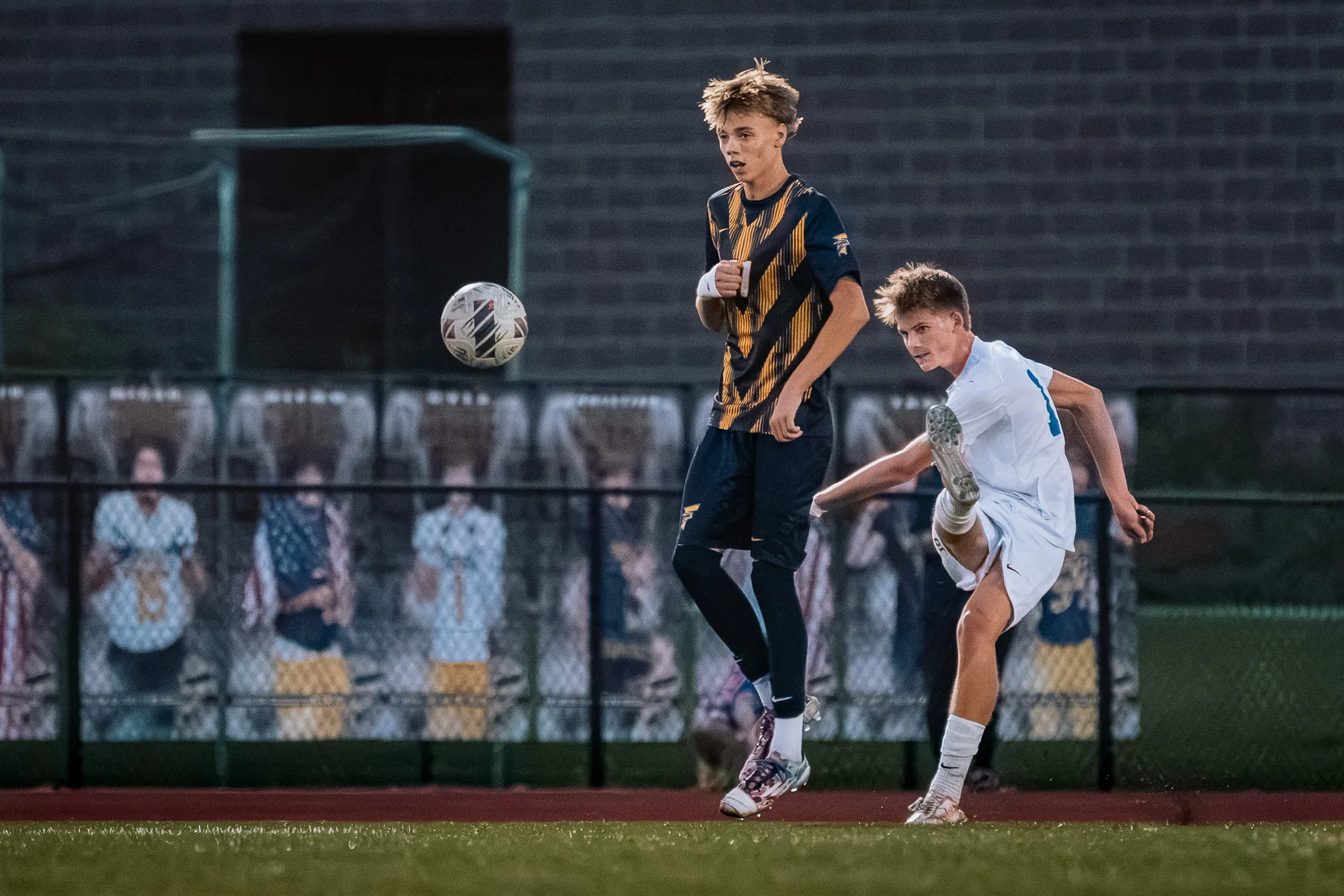 A Centaurus player swings his leg through a kick as the ball flies off to the left, a Frederick player watching beside him and printed fan banners lining the fence behind