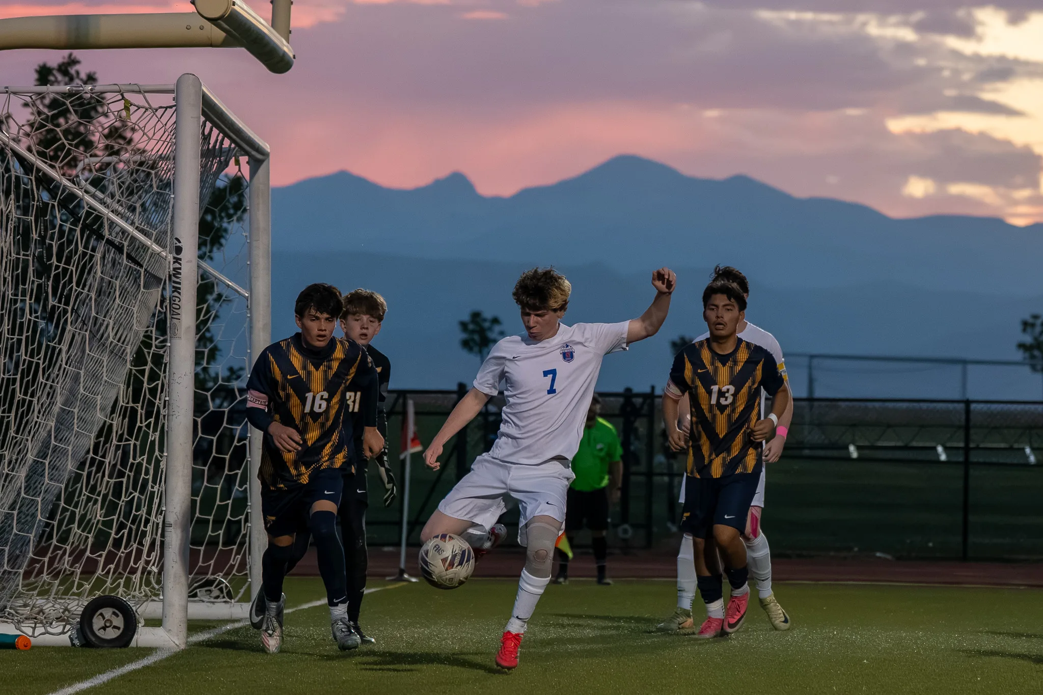 Under a pink sky and the mountains, Centaurus number 7 plays the ball near the goal as Frederick numbers 16 and 13 and others move in, an assistant referee in green behind
