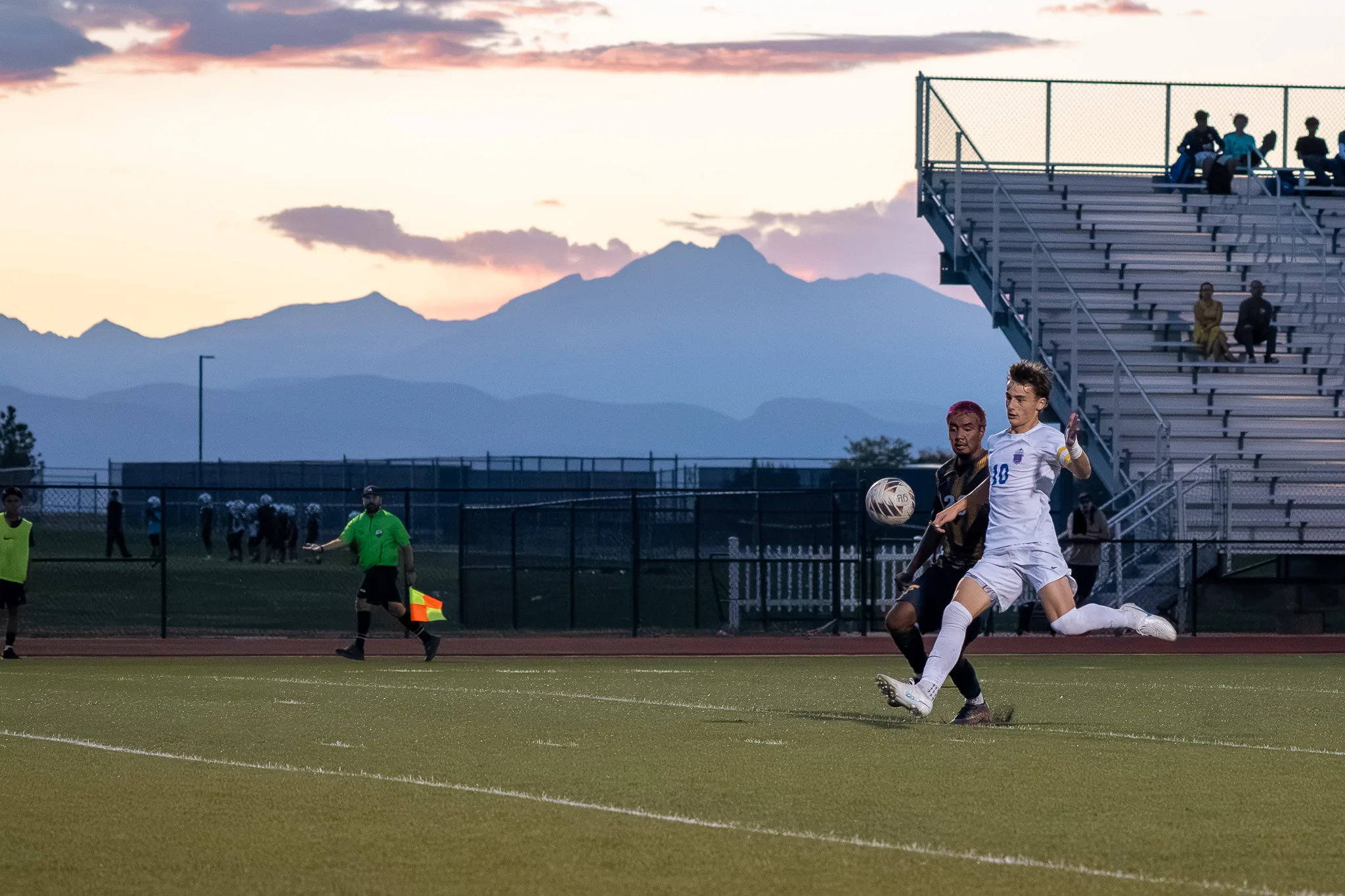 At dusk with the mountains behind them, Centaurus number 10 and a Frederick player race for the ball as an assistant referee in green runs the sideline