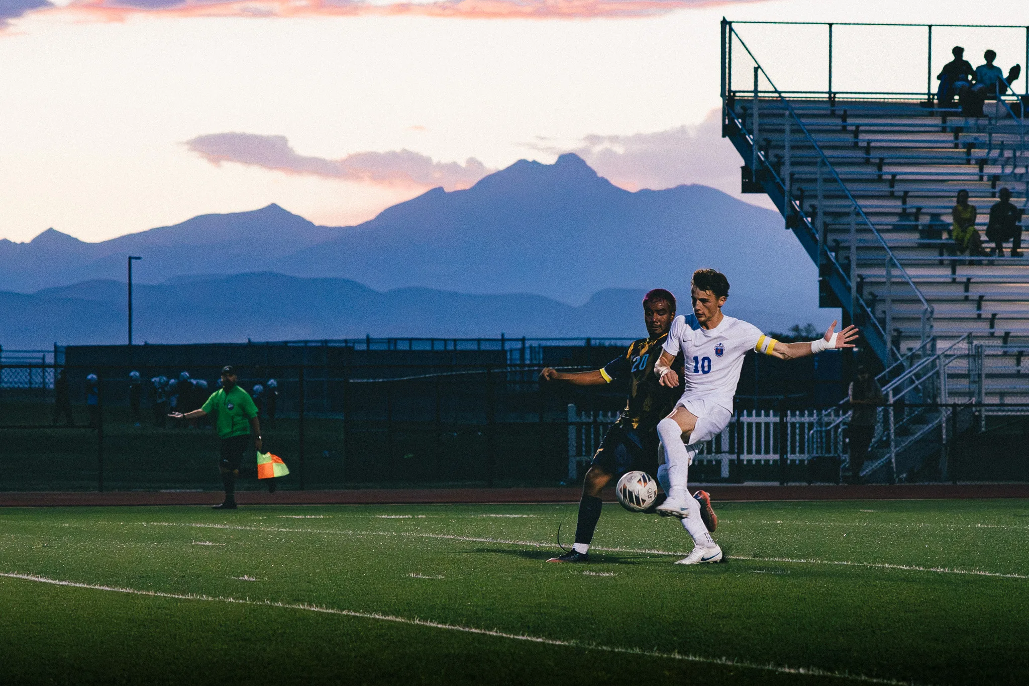 Centaurus number 10 stretches a leg to the ball as Frederick number 20 reaches in from behind, the mountains and bleachers in the dusk beyond