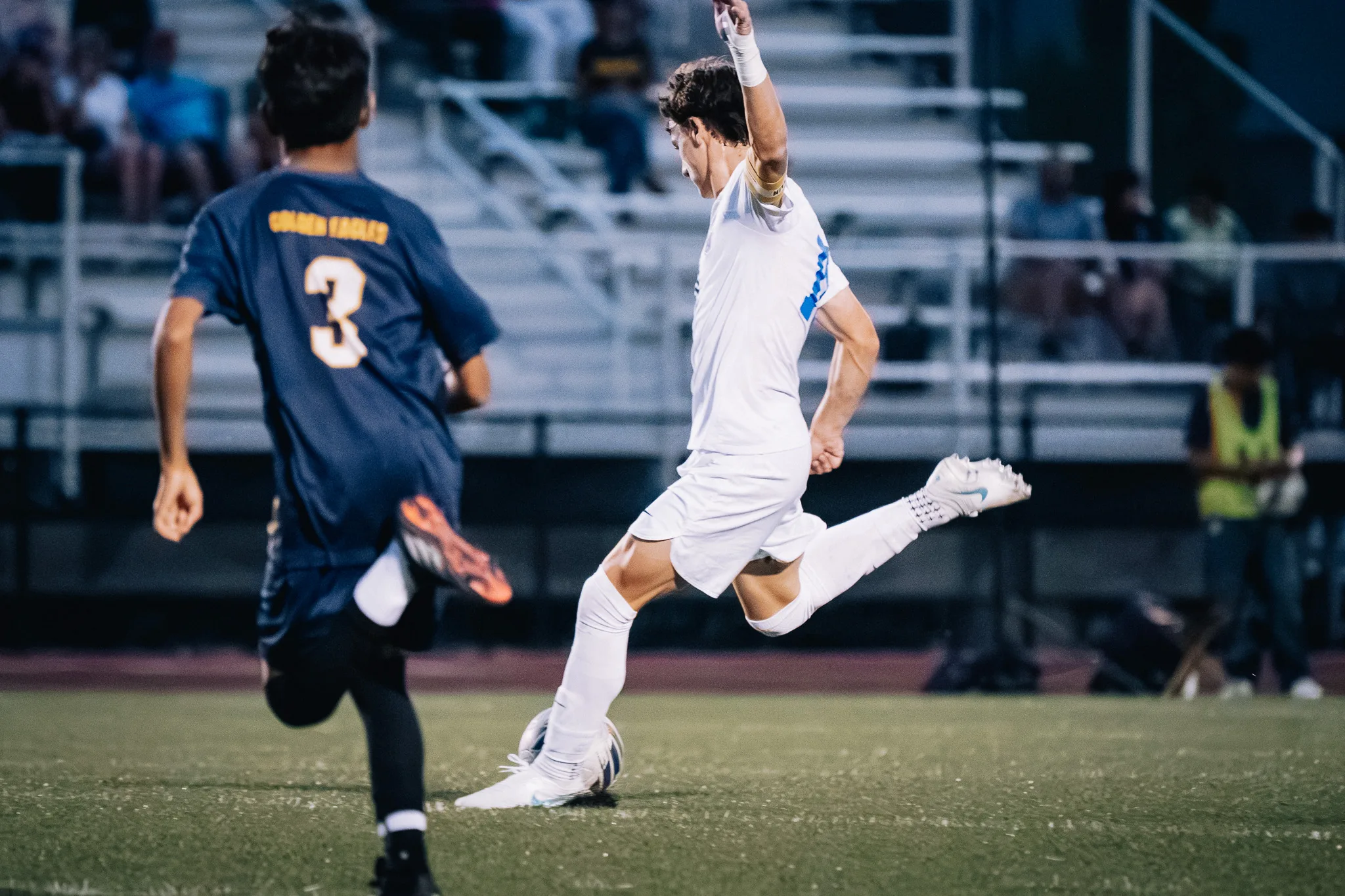 A Centaurus player in a yellow armband swings his leg through the ball with one arm raised as Frederick number 3 runs past in the foreground
