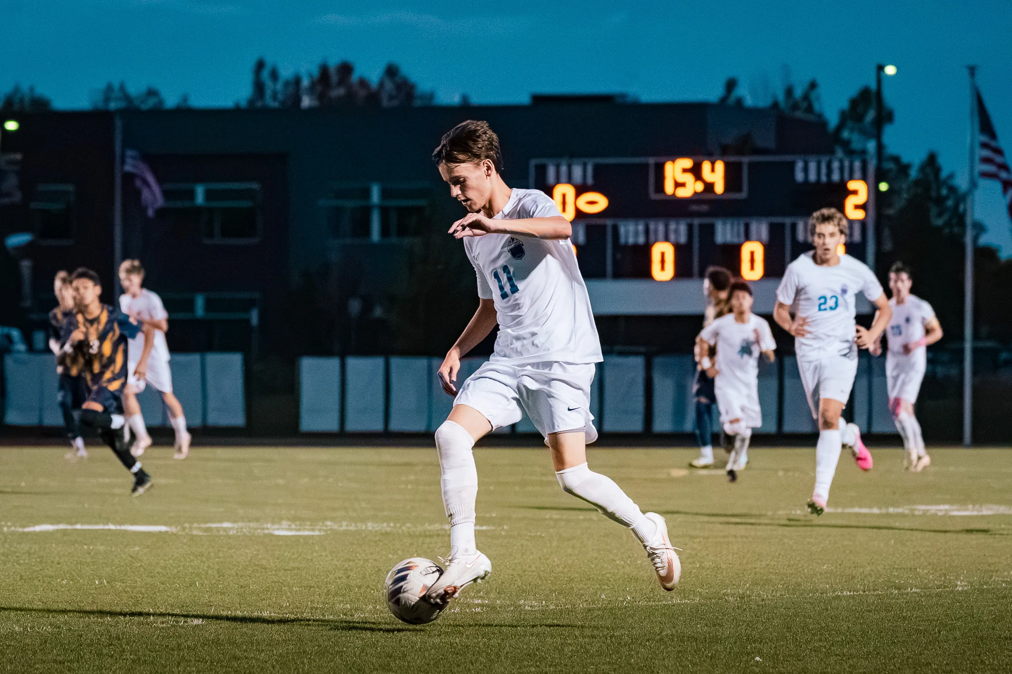 Centaurus number 11 dribbles the ball under the scoreboard in the blue evening light as teammates, including number 23, run up the field behind him