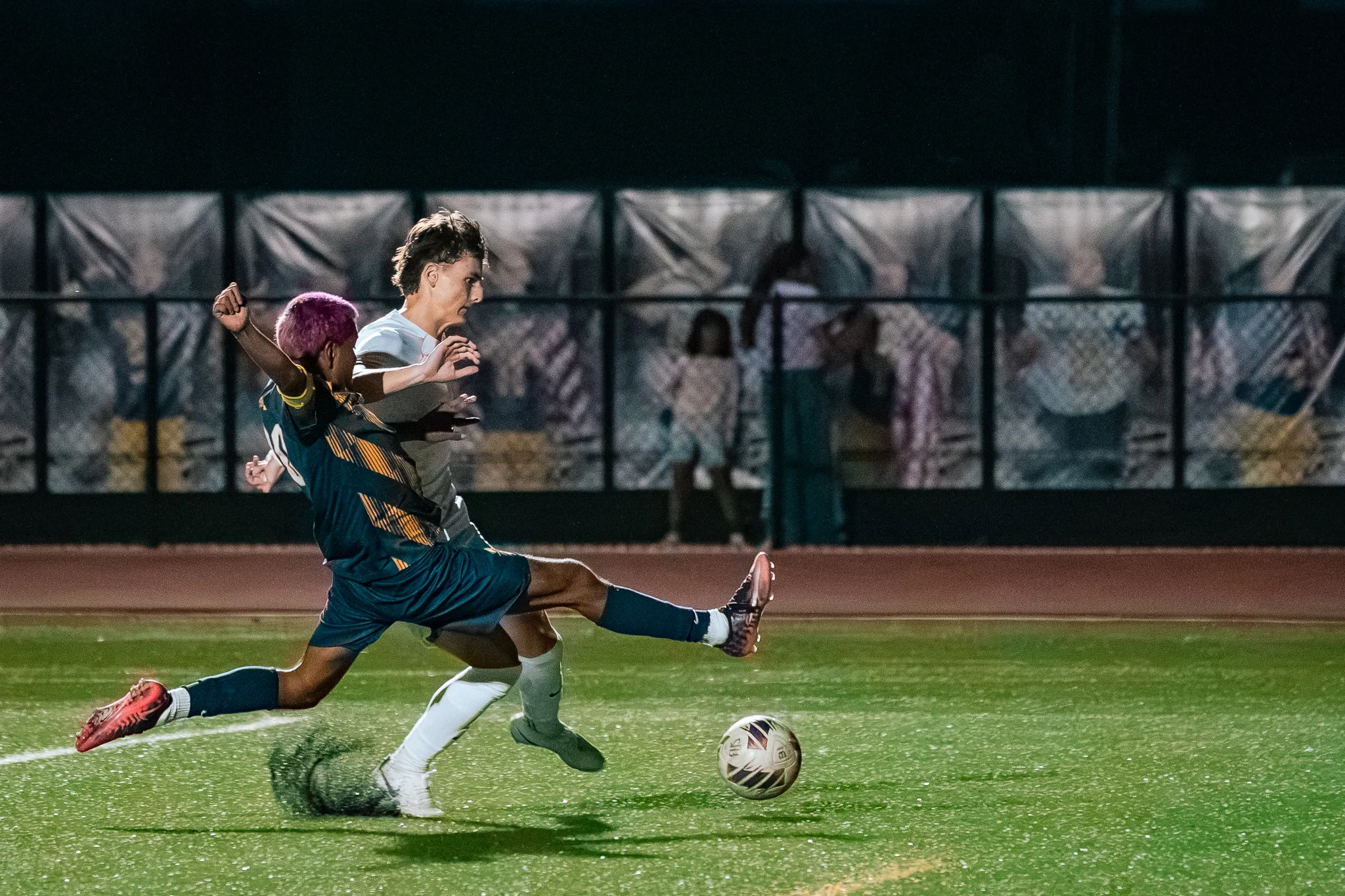 A Frederick player with pink hair stretches a leg toward the ball as a Centaurus player beside him raises both arms, spectators behind the fence