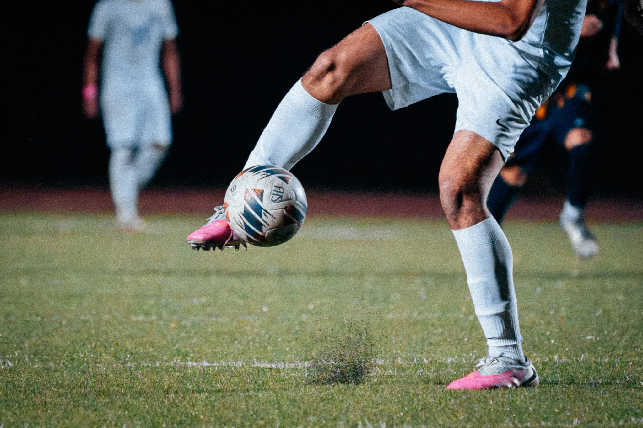 In pink cleats, a Centaurus player kicks the ball with specks of turf hanging in the air below it, a teammate and a Frederick player blurred behind