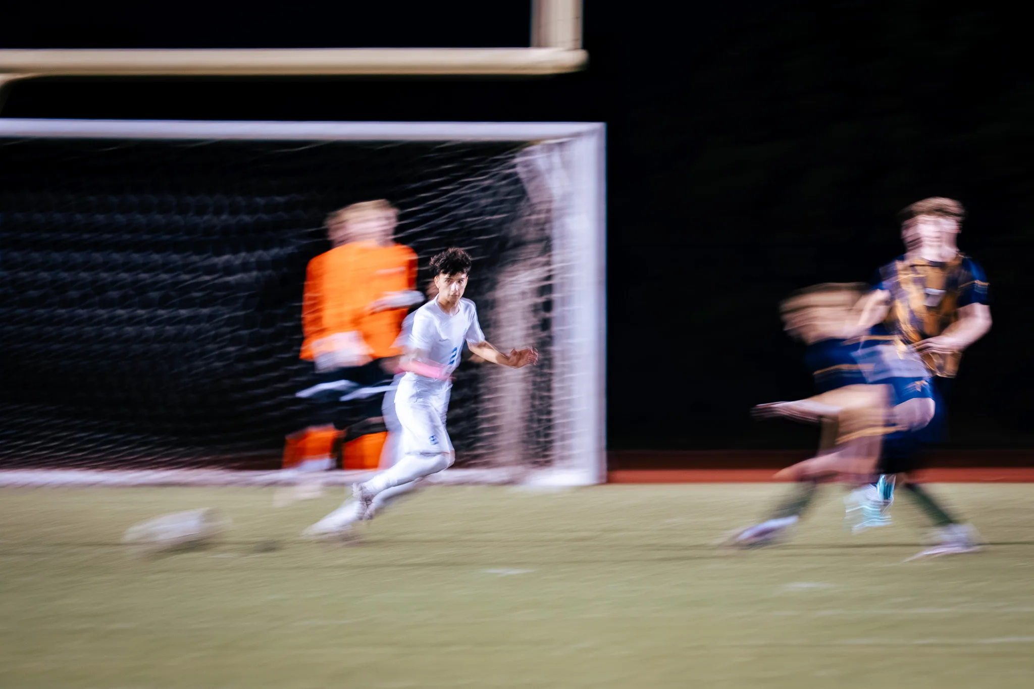 A Centaurus player runs across the front of the goal in a blur, the goalkeeper in orange smeared behind him and two Frederick players streaking through on the right