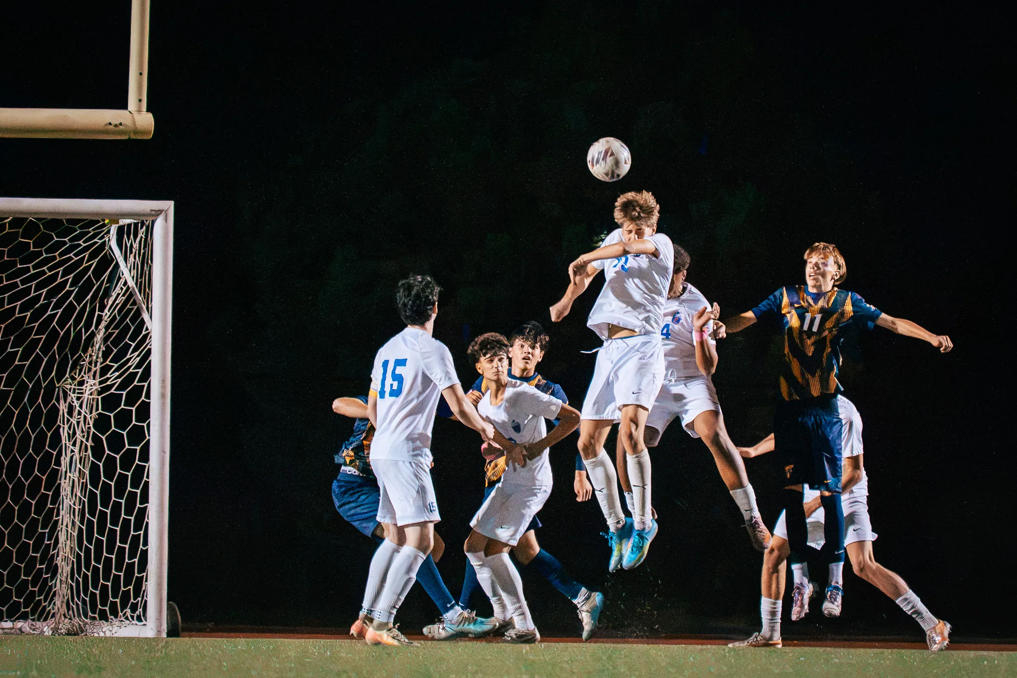 A Centaurus player rises above the crowd to head the ball in front of the goal at night, Centaurus number 15 nearby and Frederick number 11 leaning in