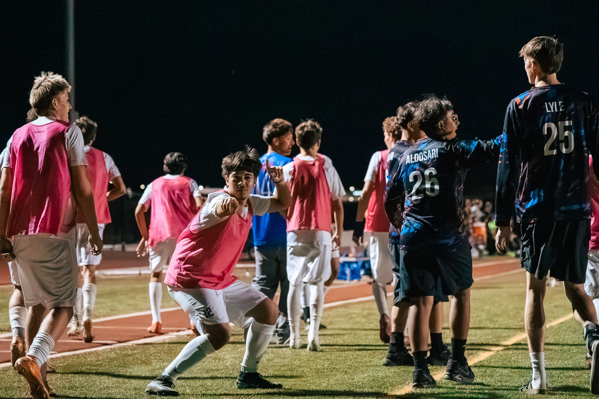 On the sideline, a Centaurus substitute in a pink bib crouches with a fist toward the camera and a hang loose sign, teammates and Frederick players walking past