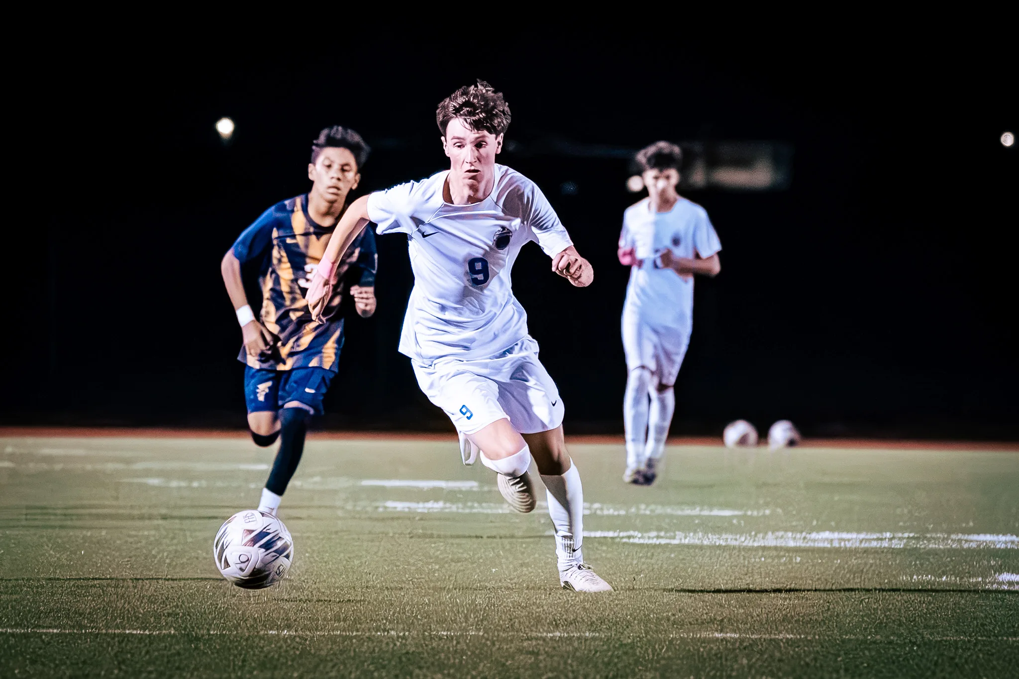 At night, Centaurus number 9 sprints with the ball at his feet, a Frederick player chasing and a Centaurus teammate jogging behind