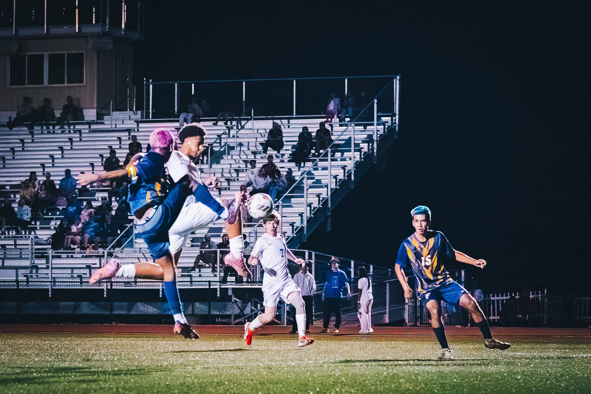 Beside the bleachers, a Frederick player and a Centaurus player both swing a leg at the ball in the air as Centaurus number 7 runs in and Frederick number 15 looks on