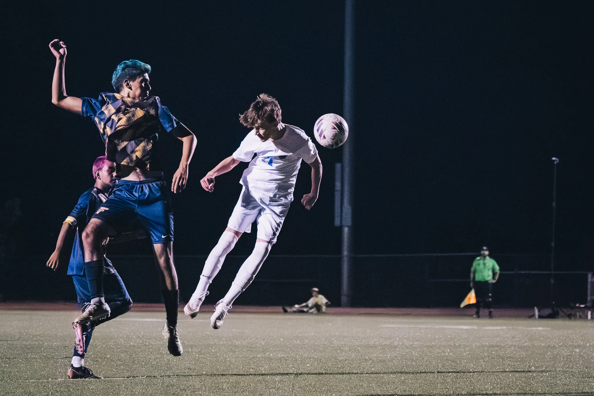 A Centaurus player stretches out flat in the air to head the ball past a Frederick player with teal hair, an assistant referee in green watching from the far side