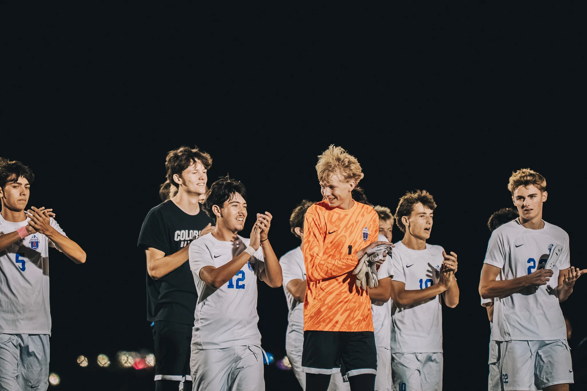 Smiling and clapping in the dark, Centaurus players, including numbers 5, 12 and 10, gather around the goalkeeper in orange, number 1, who holds his gloves