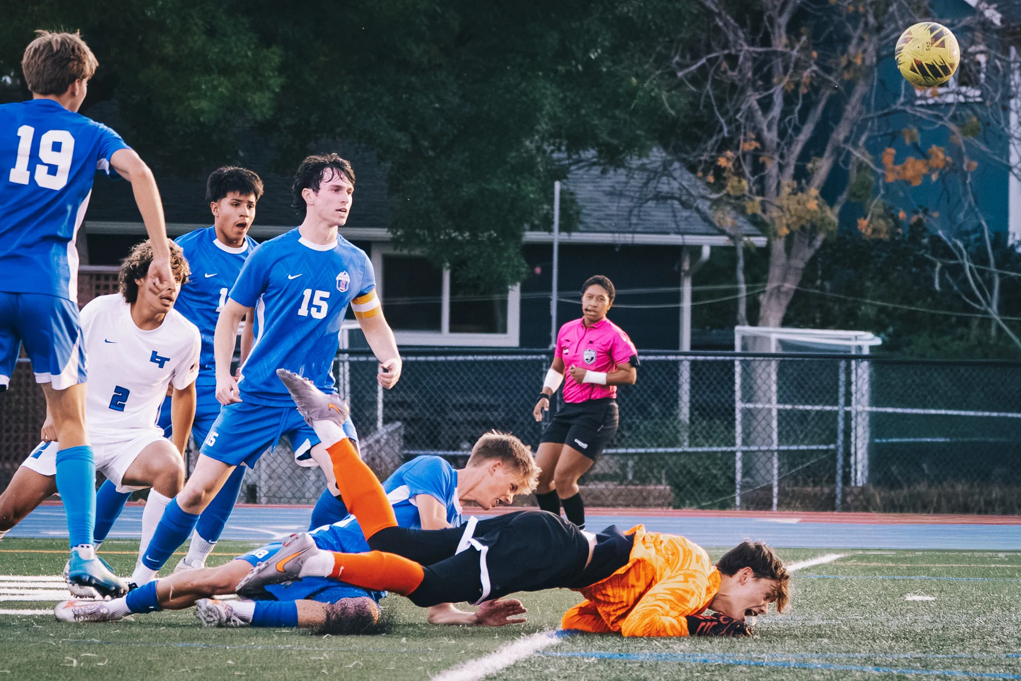 The Centaurus goalkeeper in orange sprawls on the turf beside a fallen teammate as the ball flies overhead and the referee watches