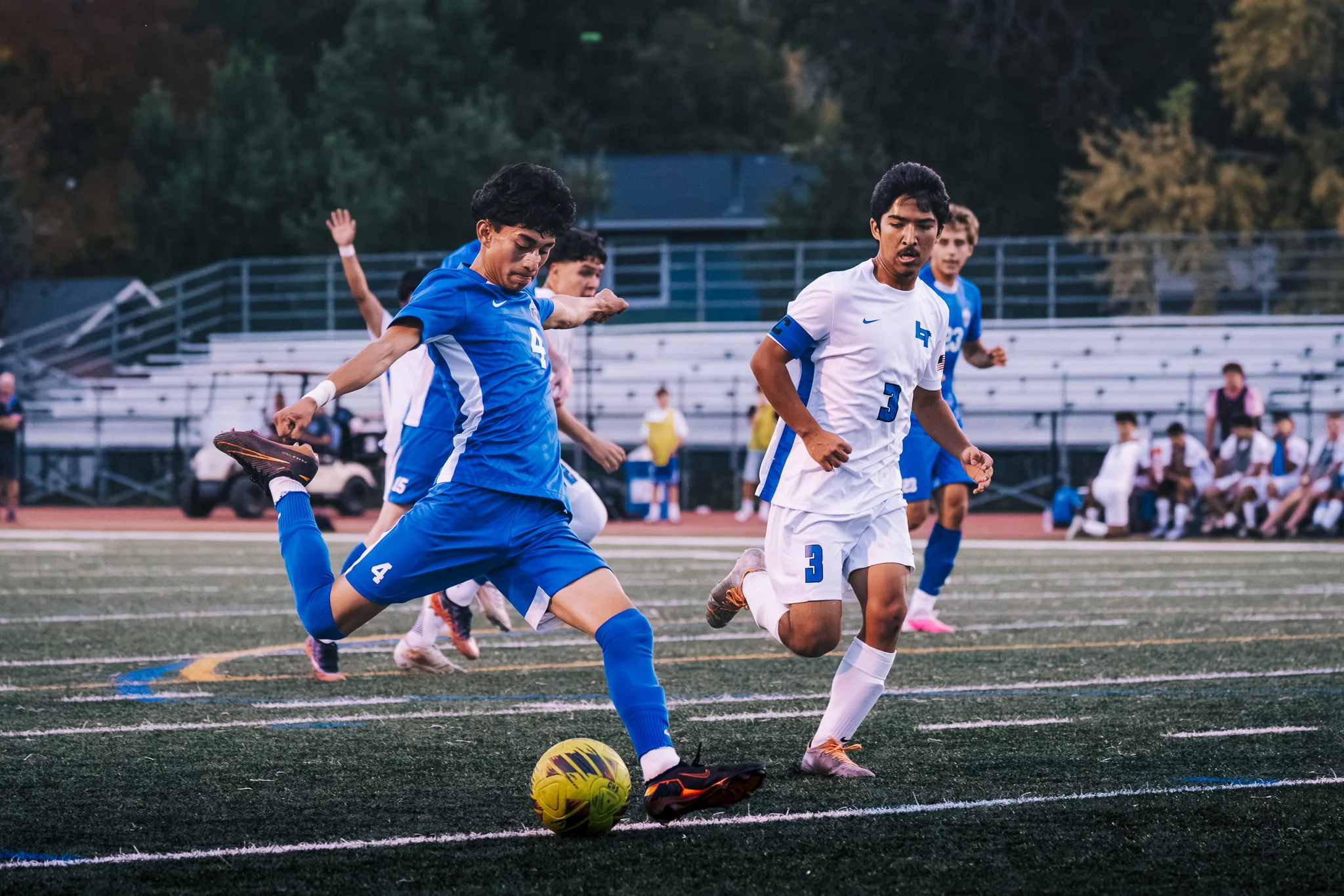 As Longmont number 3 runs in beside him, Centaurus number 4 strikes the ball, the bleachers behind them