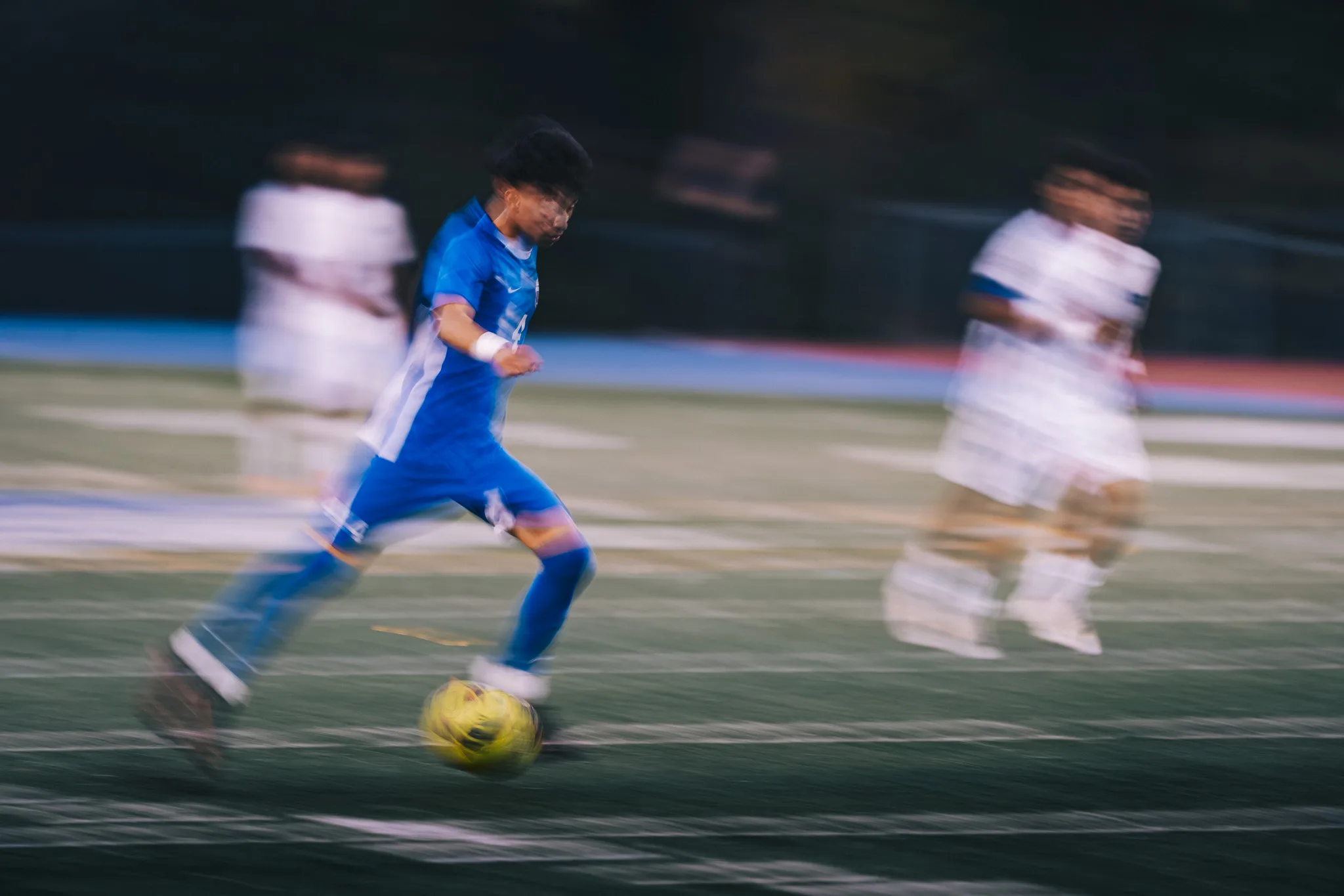 Through a streak of motion blur, a Centaurus player dribbles with a Longmont player blurred on each side