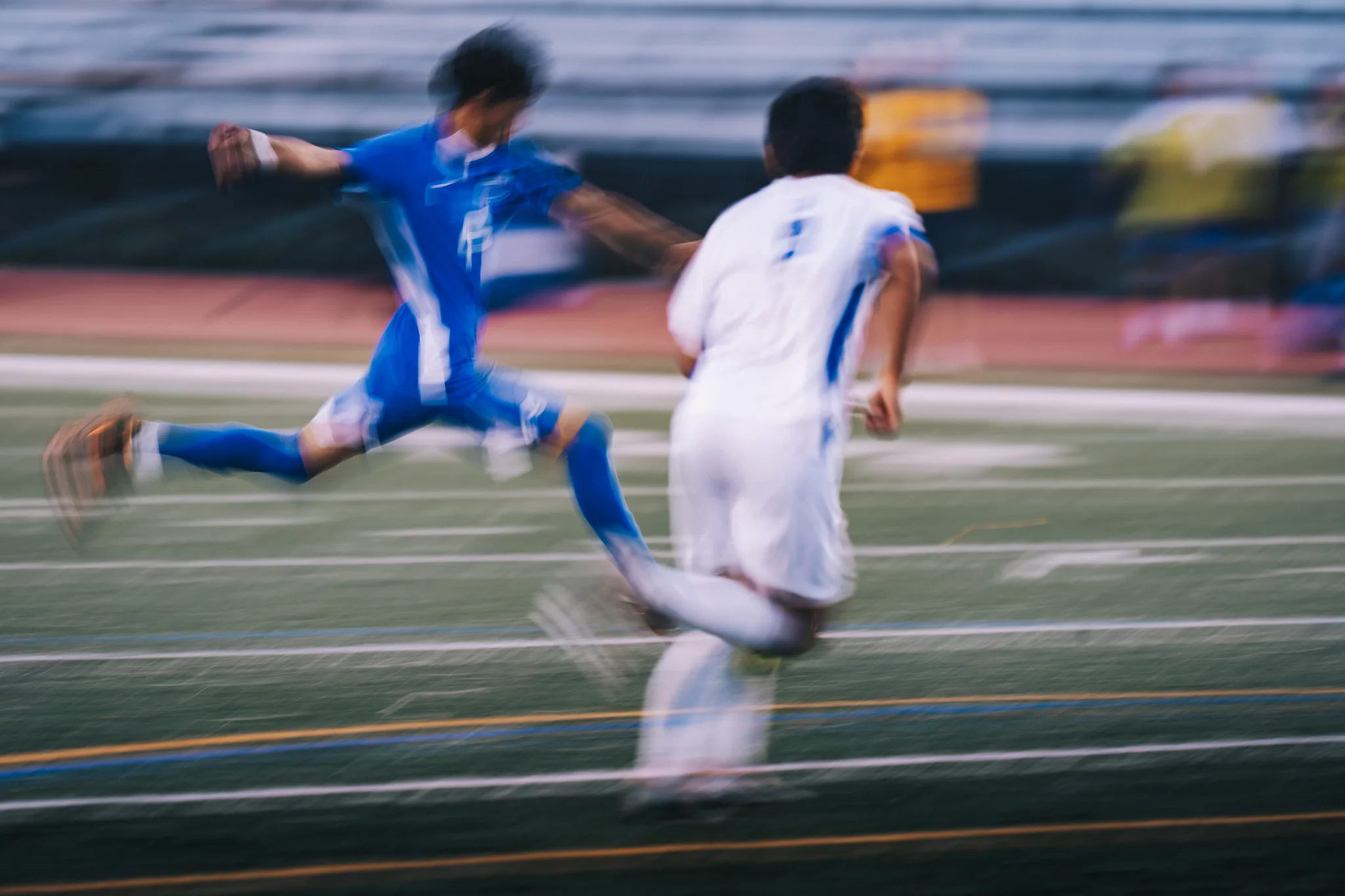 A Centaurus player stretches into a long stride beside a Longmont player in a blur of motion, the bleachers behind them
