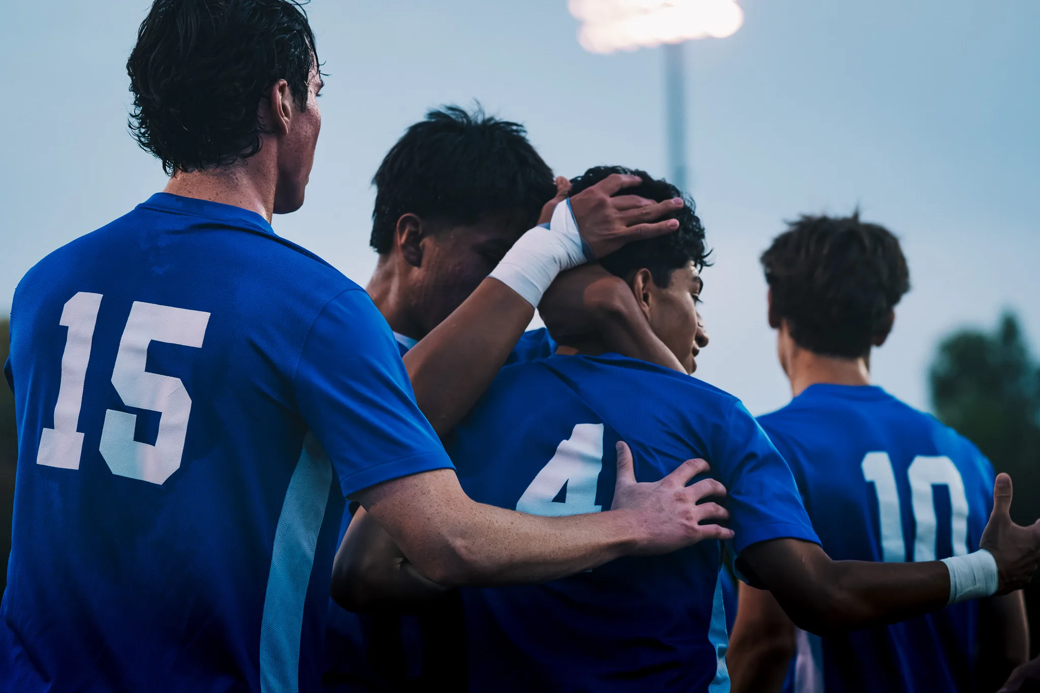 Centaurus players, including numbers 15, 4 and 10, celebrate in a tight embrace beneath a glowing stadium light