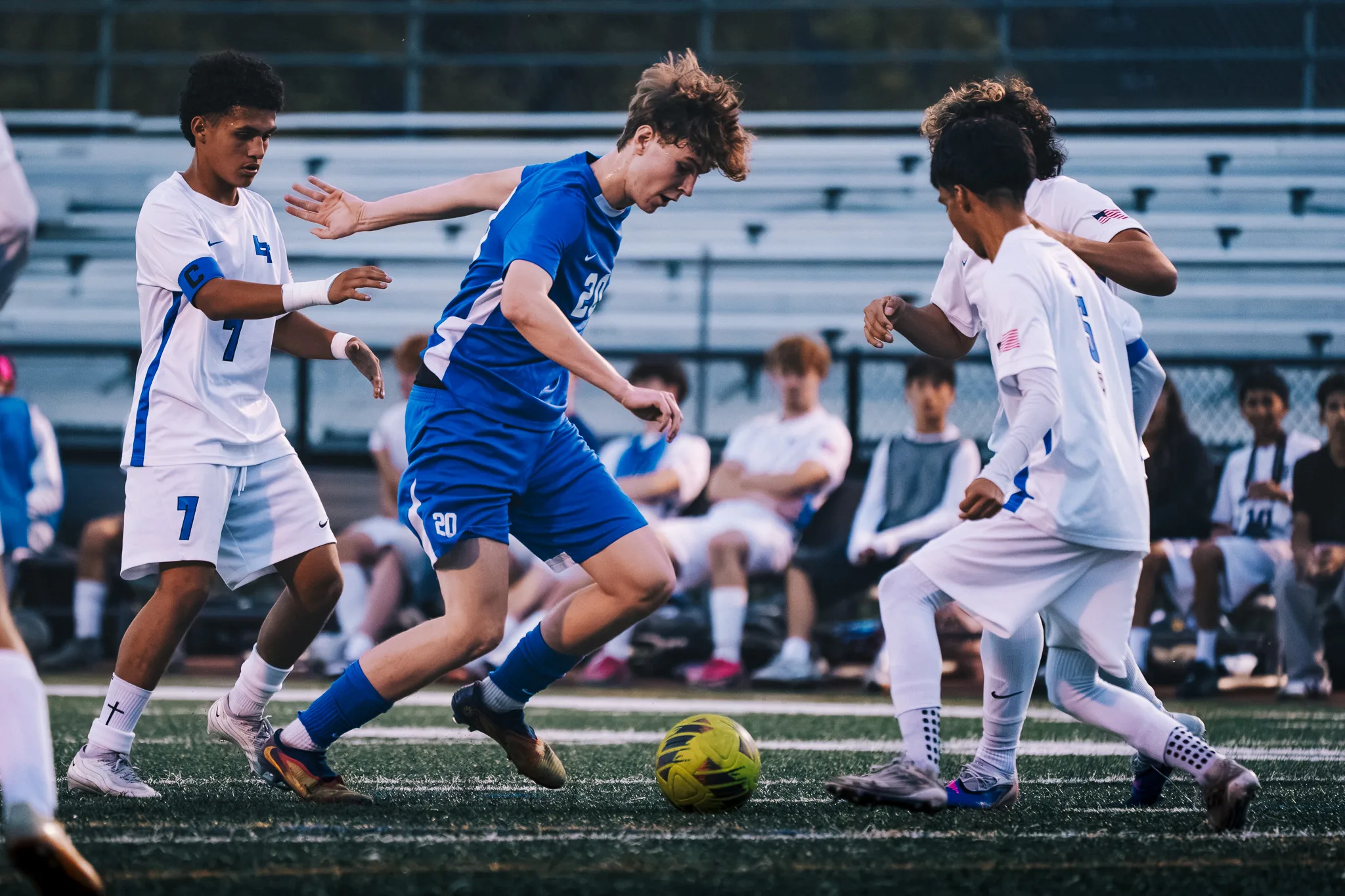 Between Longmont number 7 and two other Longmont players, Centaurus number 20 dribbles, substitutes seated behind them