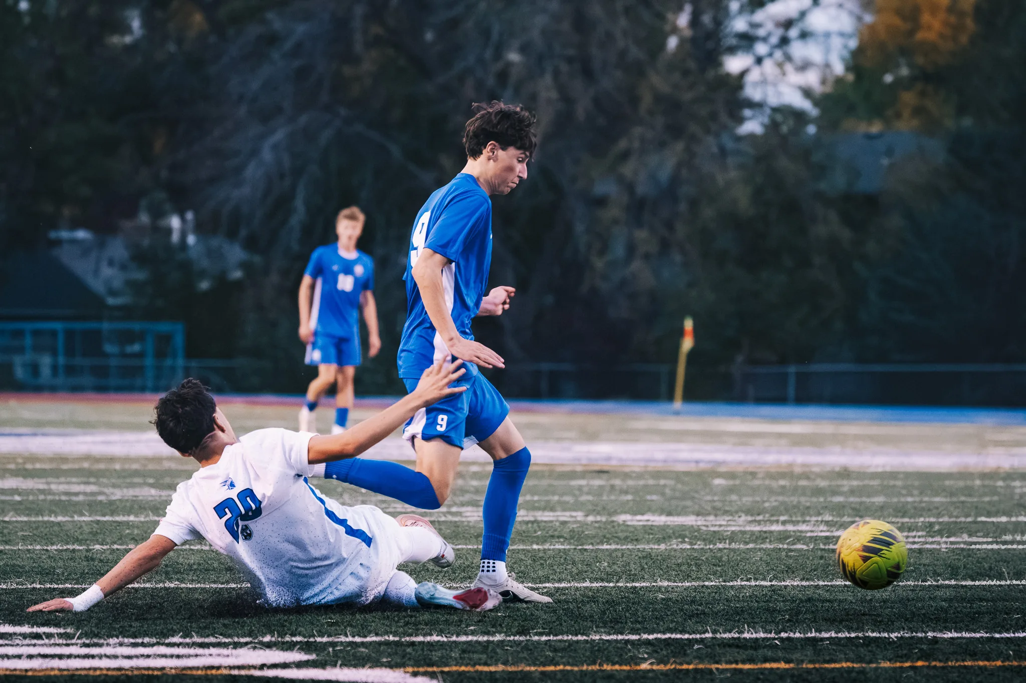A Longmont player slides across the turf as Centaurus number 9 steps over him, with Centaurus number 18 coming up behind