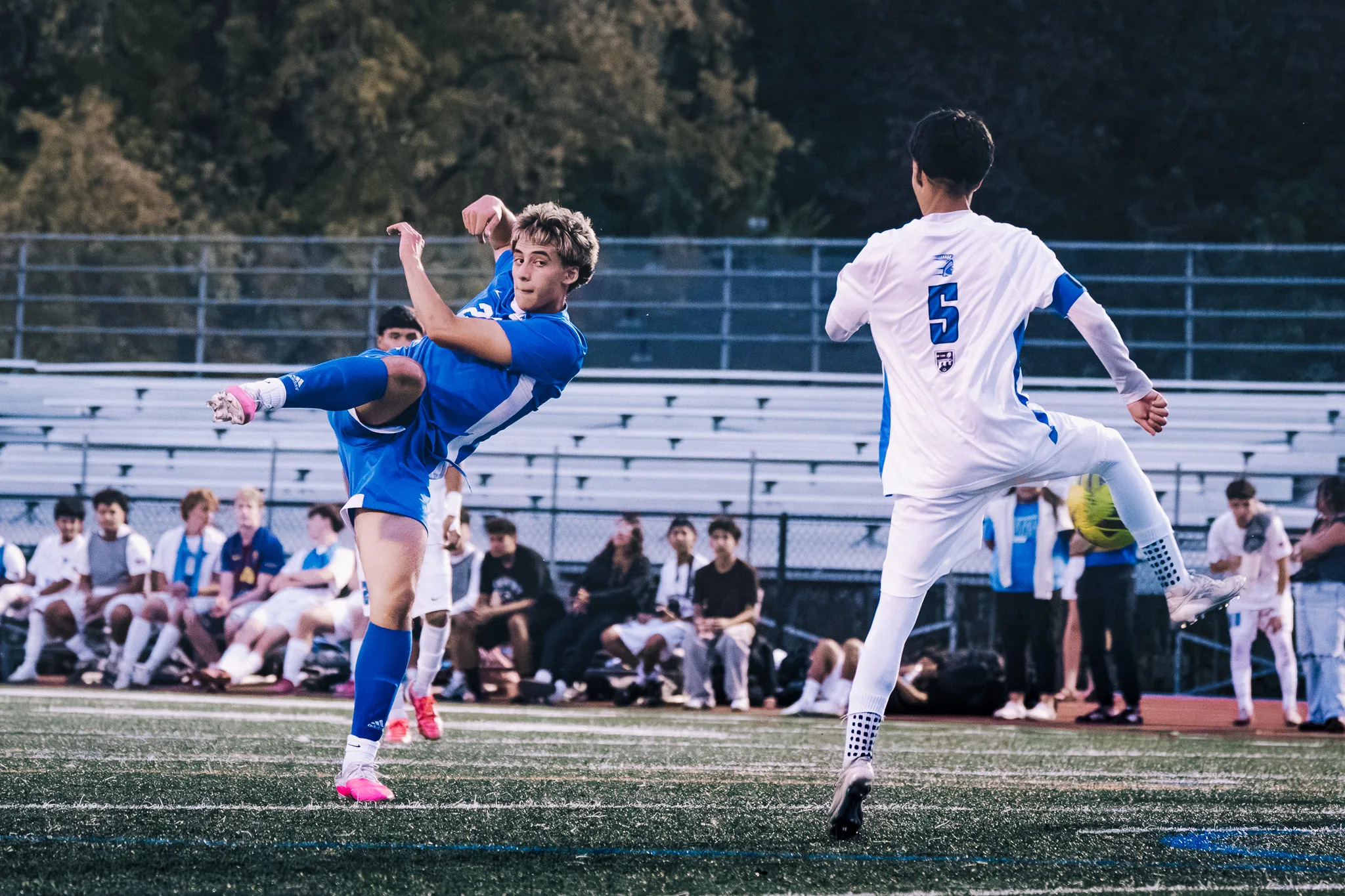 A Centaurus player leaps with one leg swung high as Longmont number 5 raises a knee beside the ball, spectators seated behind