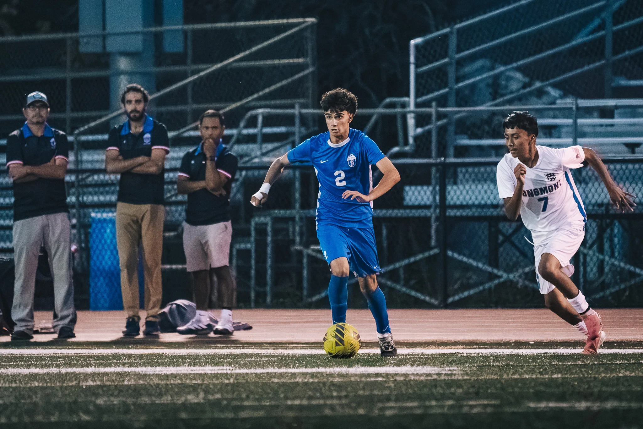Centaurus number 2 dribbles along the sideline with Longmont number 7 racing after him, three people looking on behind