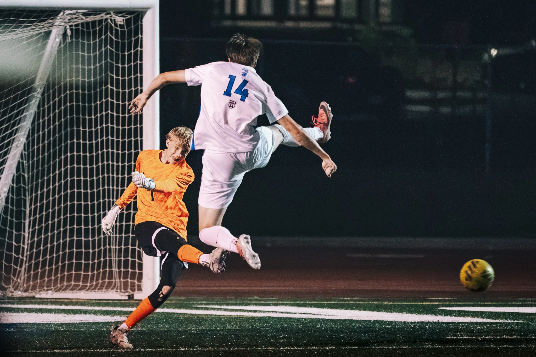 Longmont number 14 is airborne with a leg kicked up beside the Centaurus goalkeeper in orange, near the goal