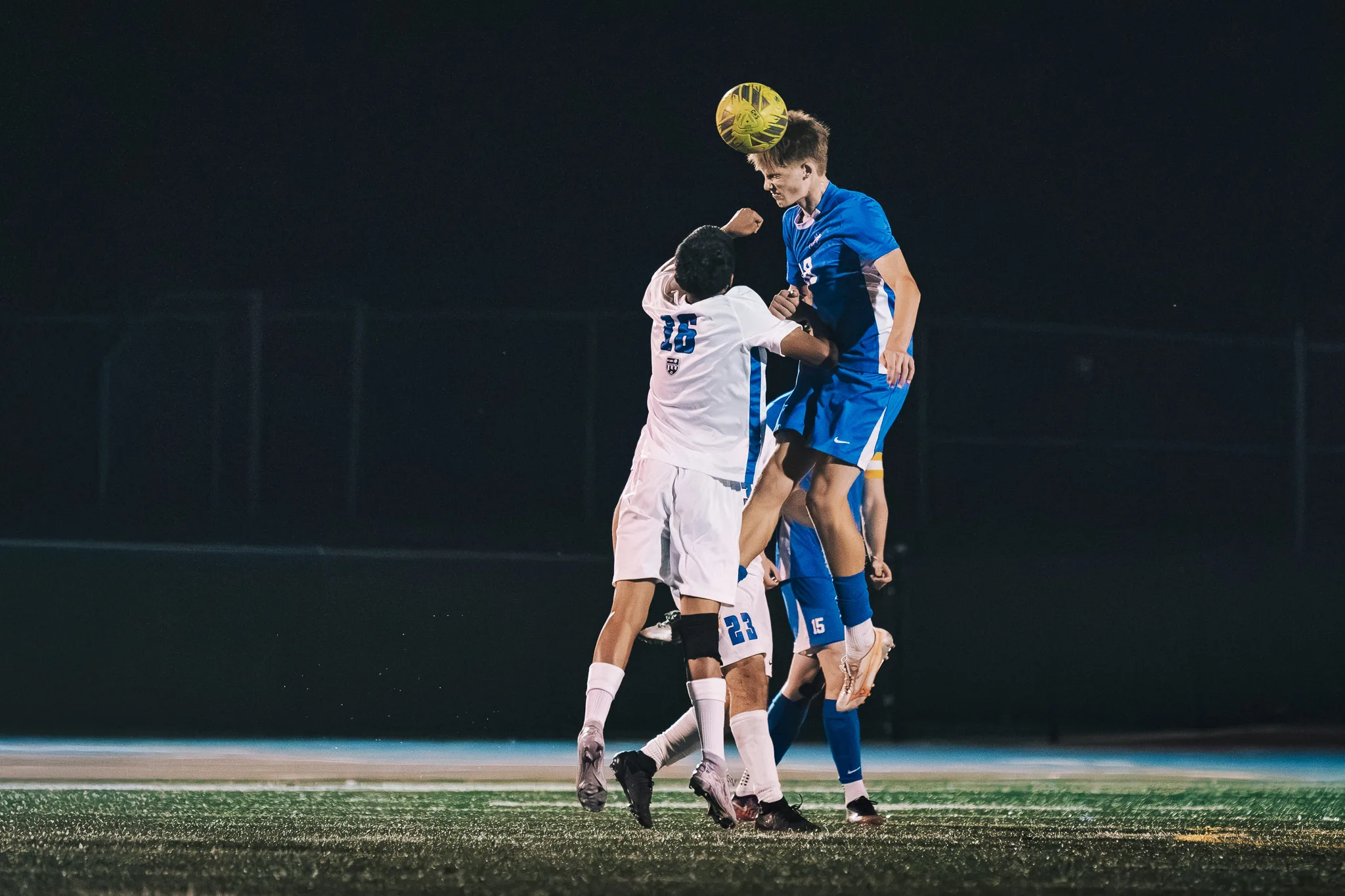 Rising above Longmont number 16, a Centaurus player heads the ball with the field fading into darkness behind them