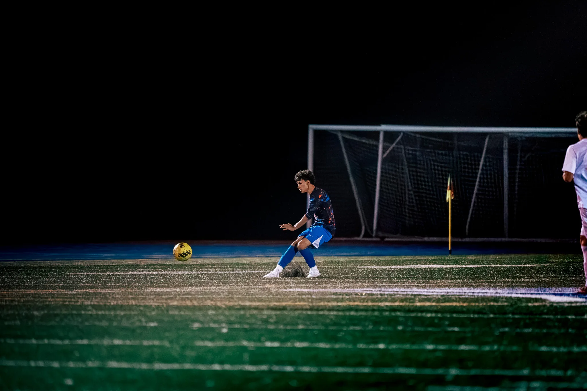 A Centaurus player follows through on a kick as the ball bounces ahead of him near a goal and a corner flag, with a Mead player entering at the right edge