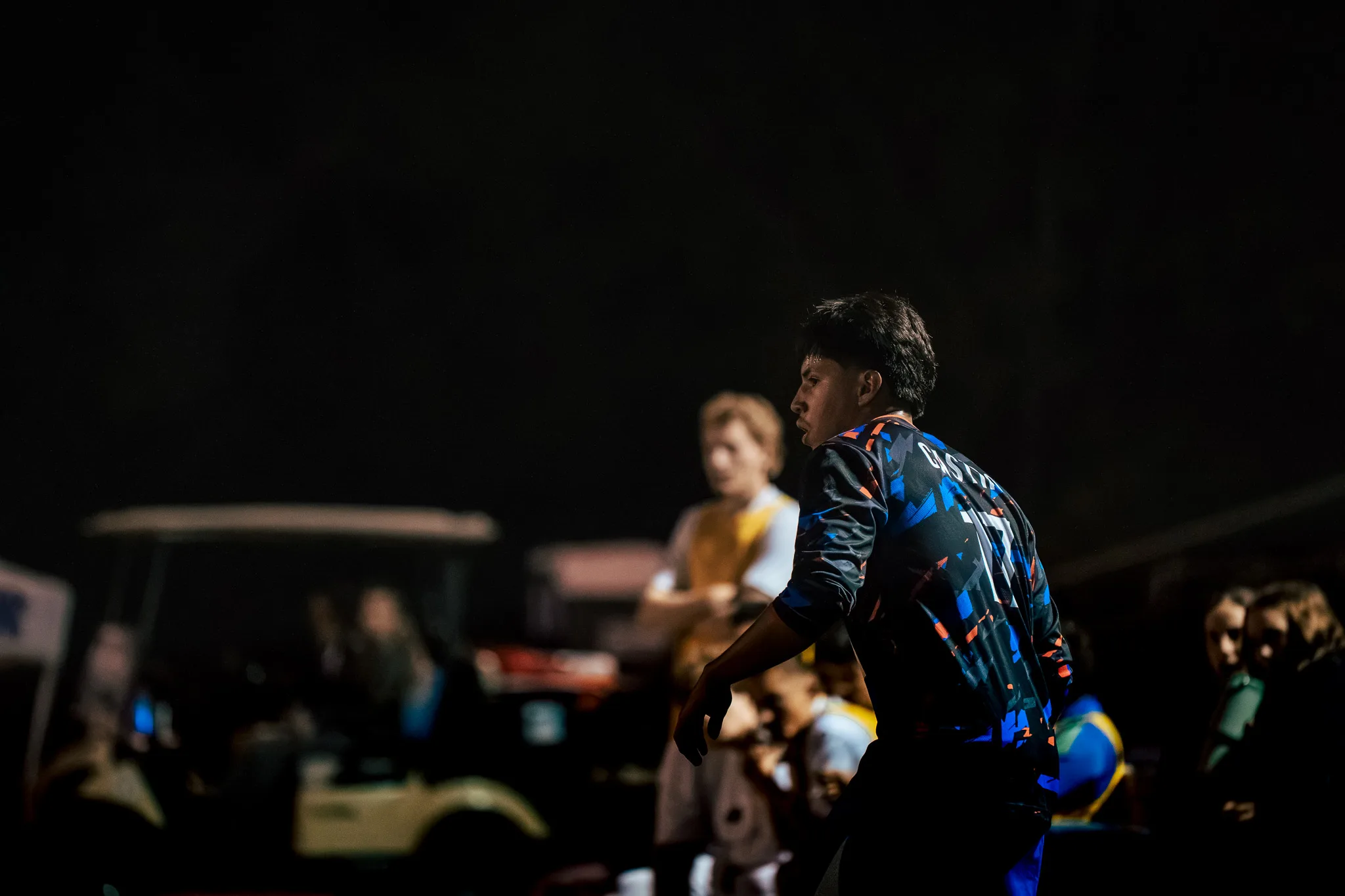 In profile, a Centaurus player walks along the sideline, a golf cart and blurred teammates and spectators behind him in the dark