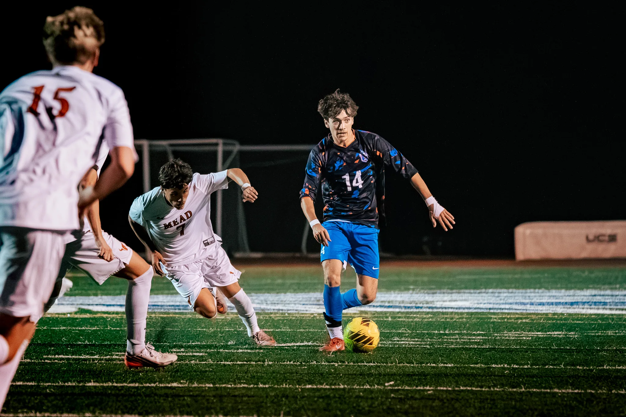 Centaurus number 14 dribbles up the field at night as Mead number 7 chases him and Mead number 15 runs across the near side